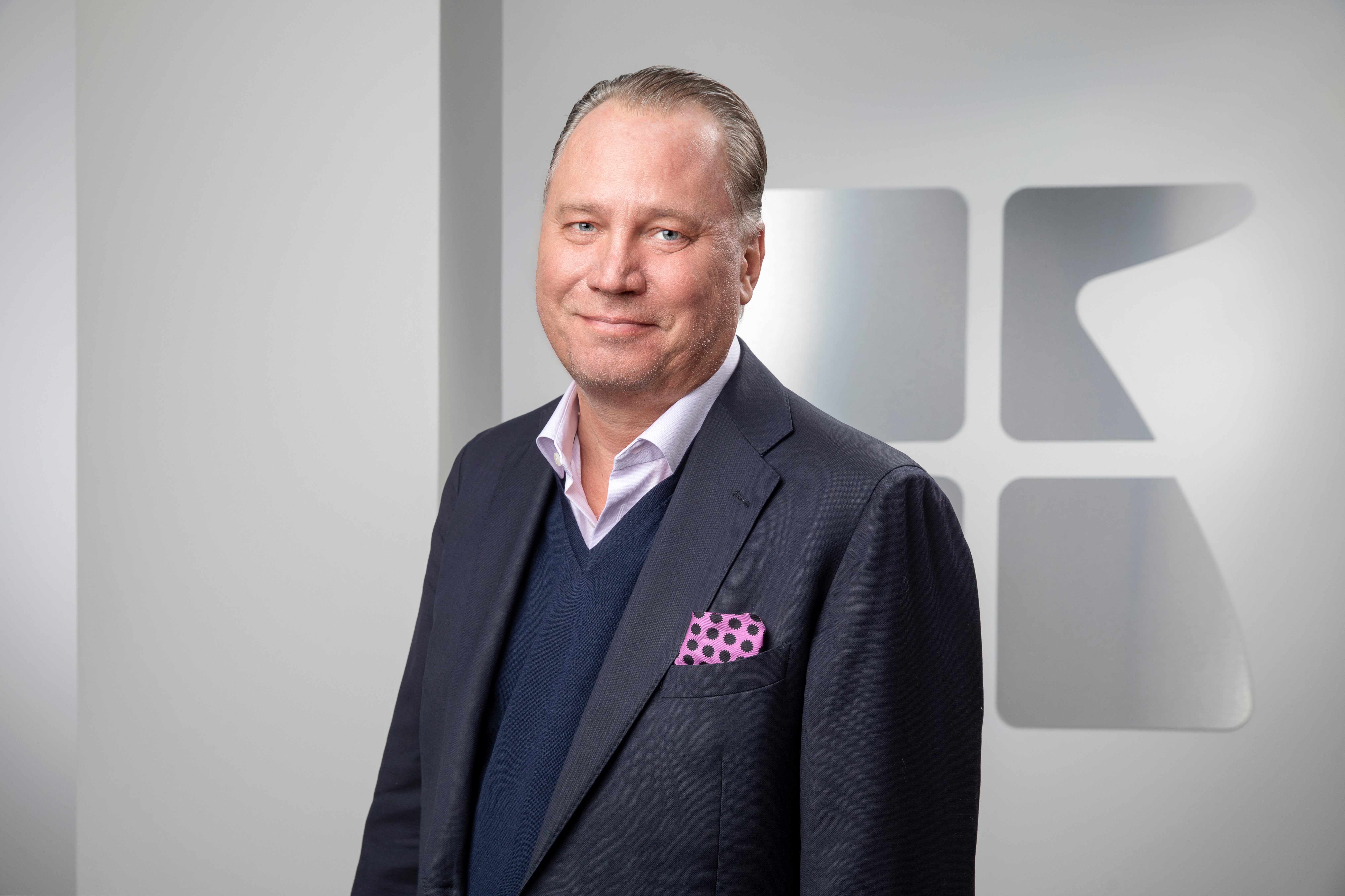 Porträtt av en man i mörk kostym och blå tröja framför en vit vägg med logotyp.