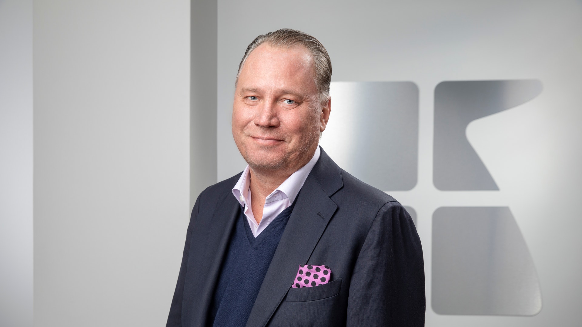 Porträtt av en man i mörk kostym och blå tröja framför en vit vägg med logotyp.