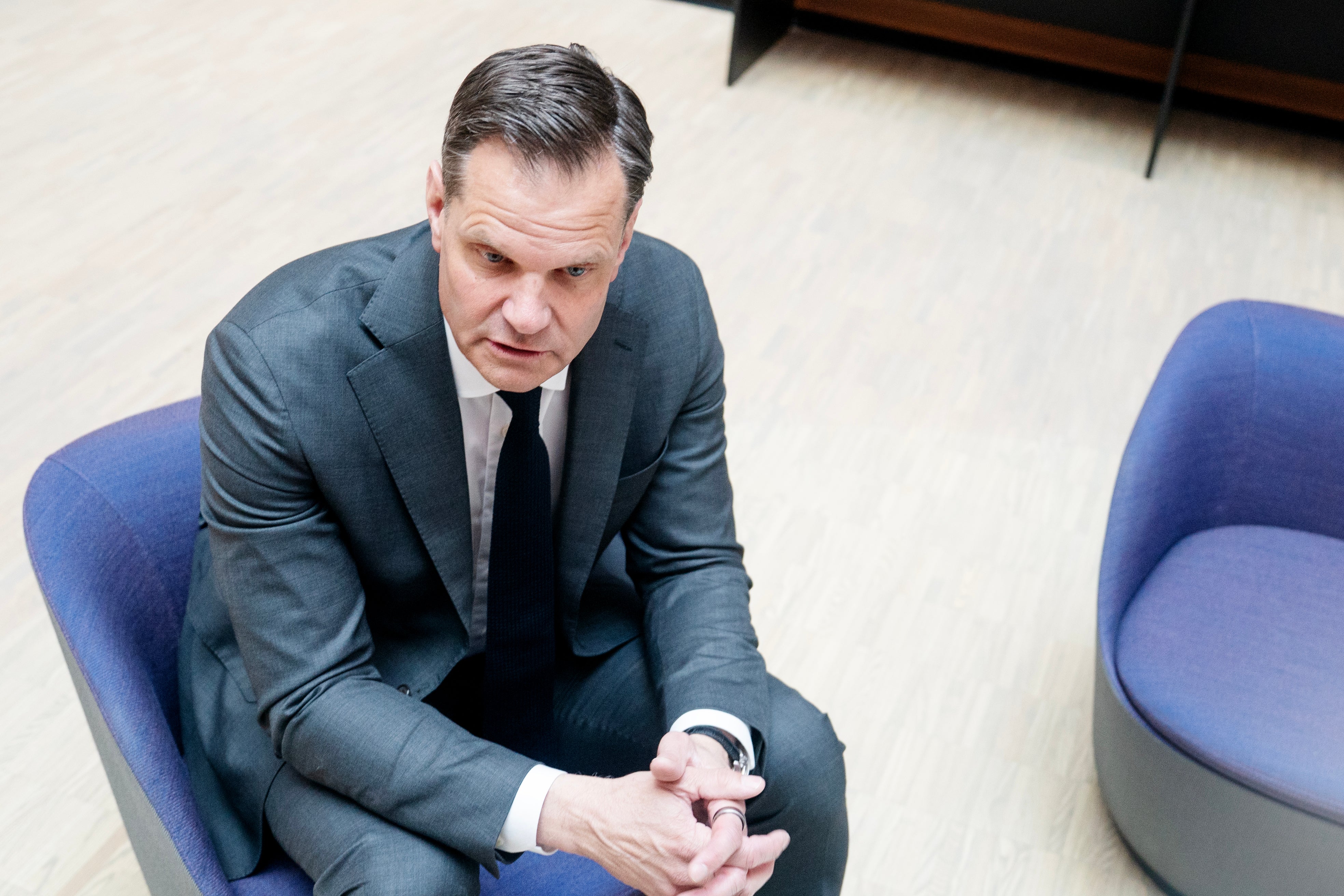 Man i kostym sitter på blå stol och tittar ner mot sina händer.