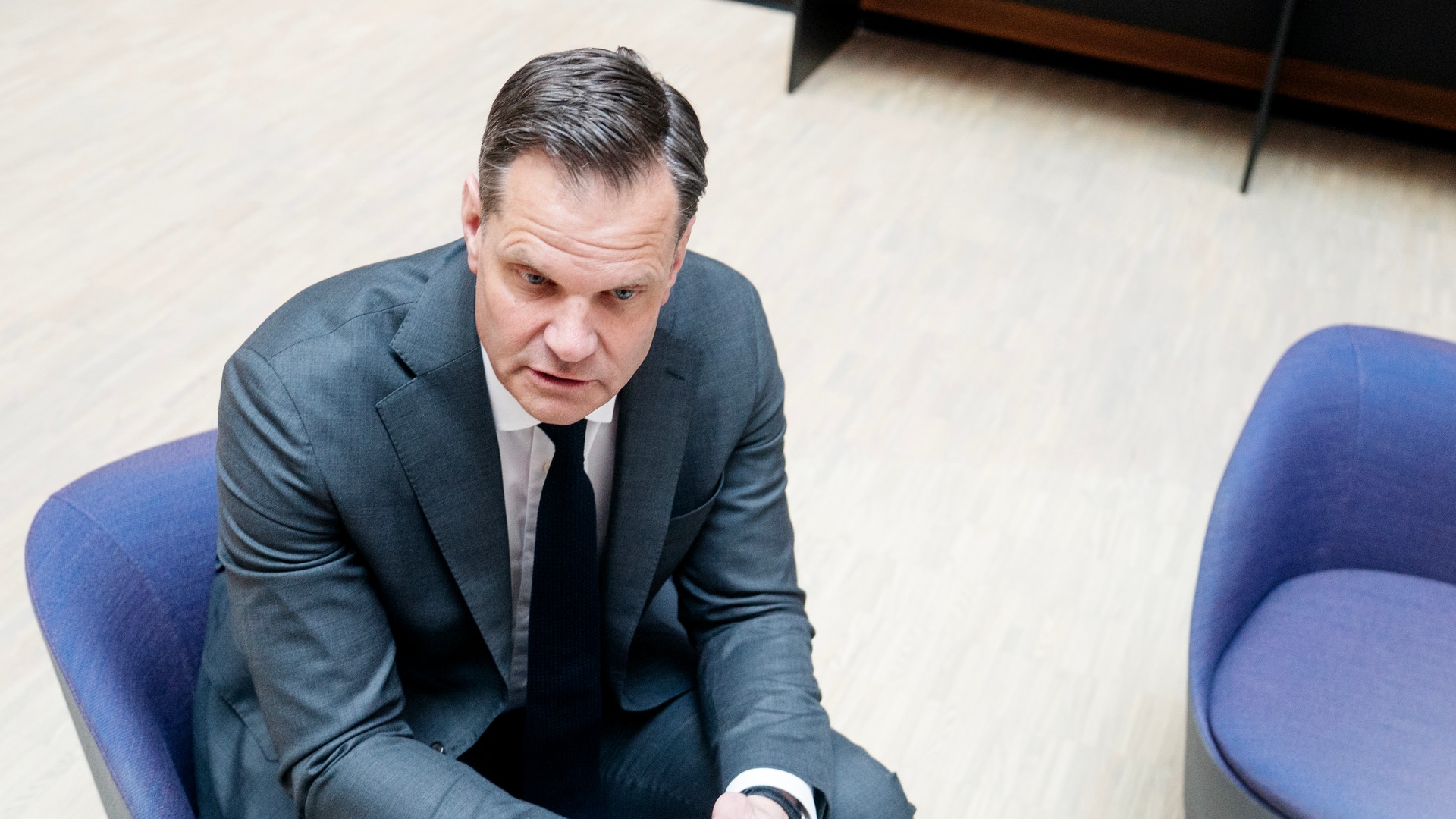 Man i kostym sitter på blå stol och tittar ner mot sina händer.