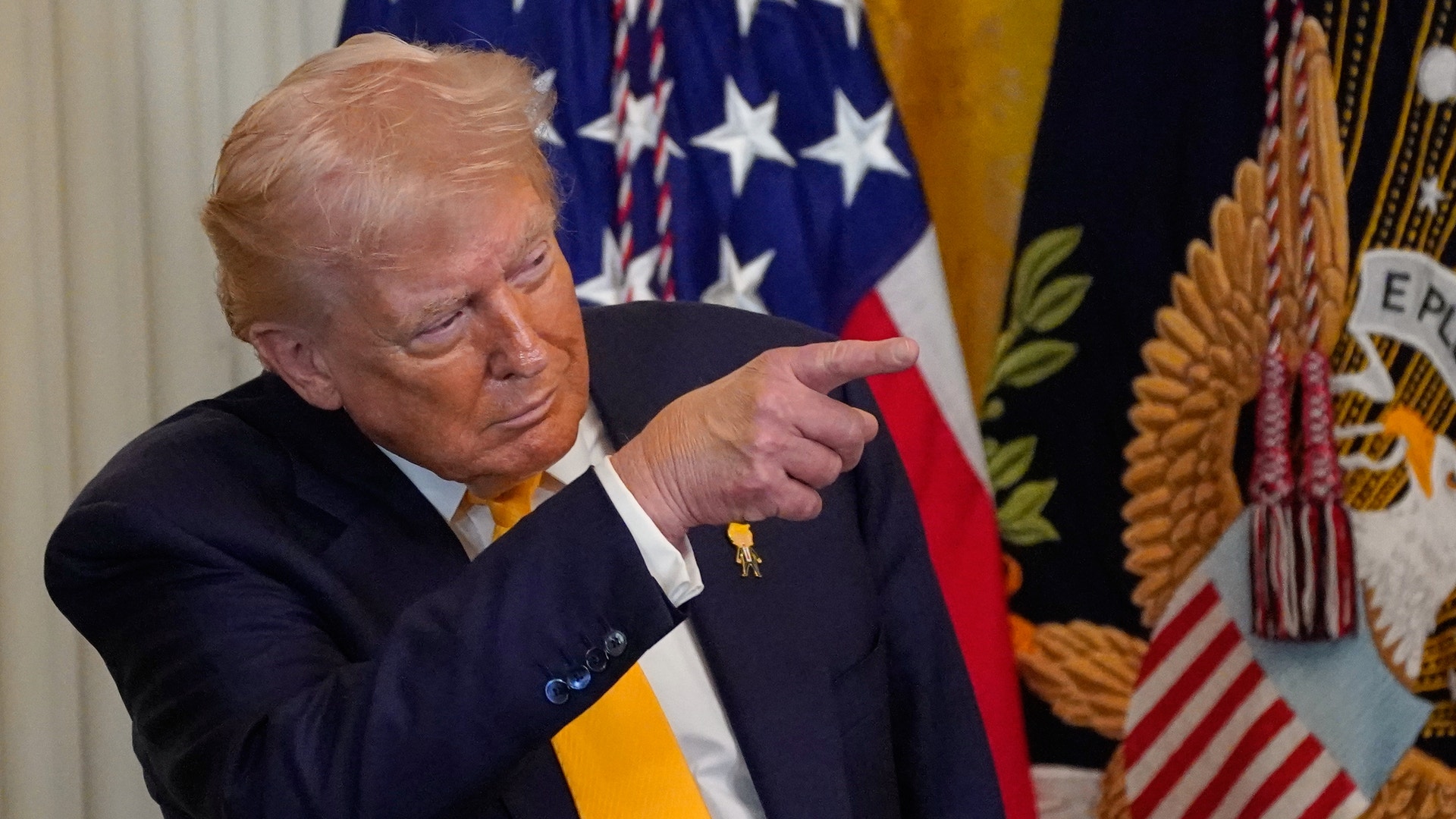 Man i mörk kostym pekar mot kameran framför amerikanska flaggor och officiella emblem.