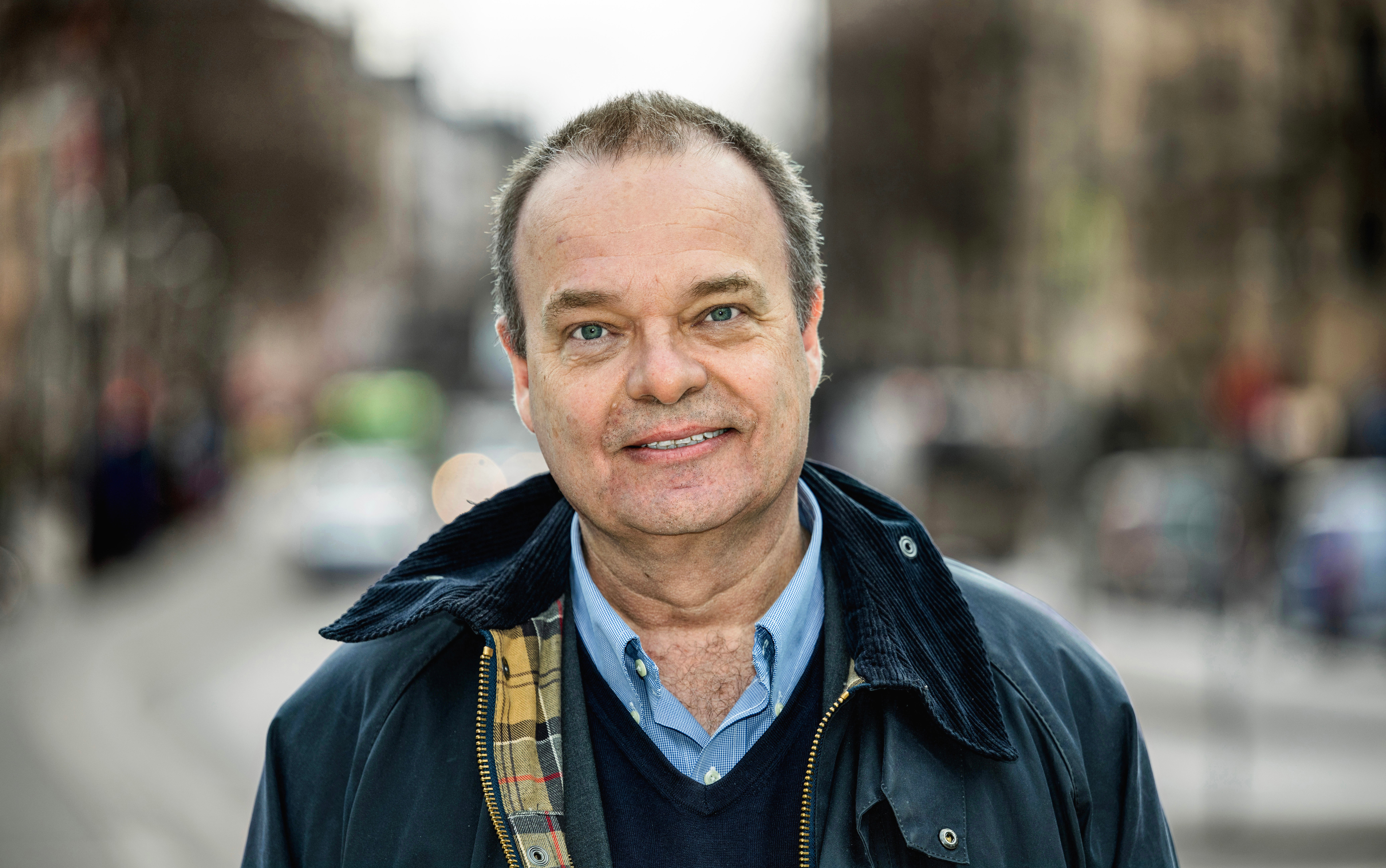 Porträtt av en 60-årig Sven Otto Littorin som ler mot kameran på en stadsgata. Han bär en mörk jacka och ljusblå skjorta.