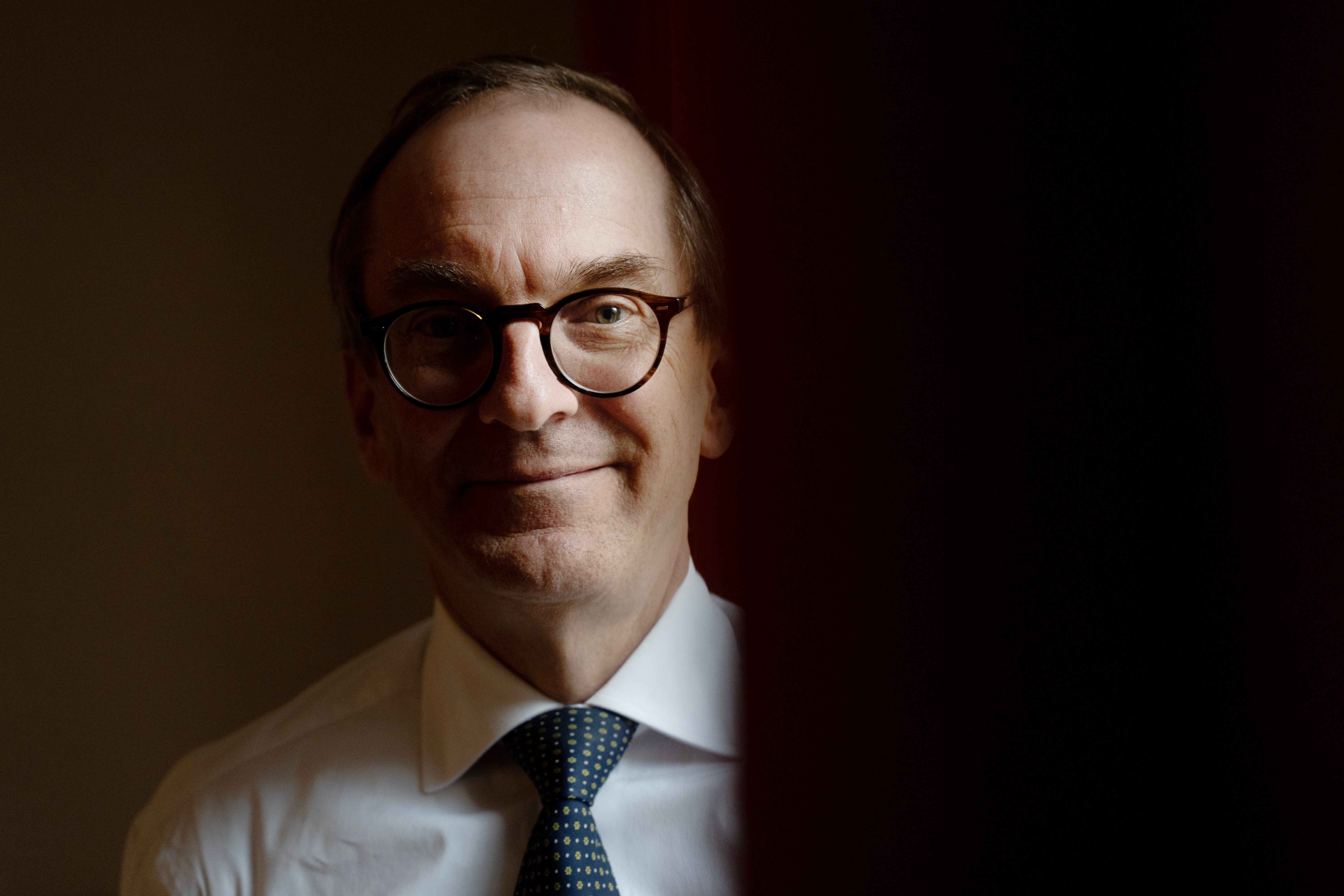 Porträtt av en man med glasögon, vit skjorta och mörkblå slips mot mörk bakgrund.