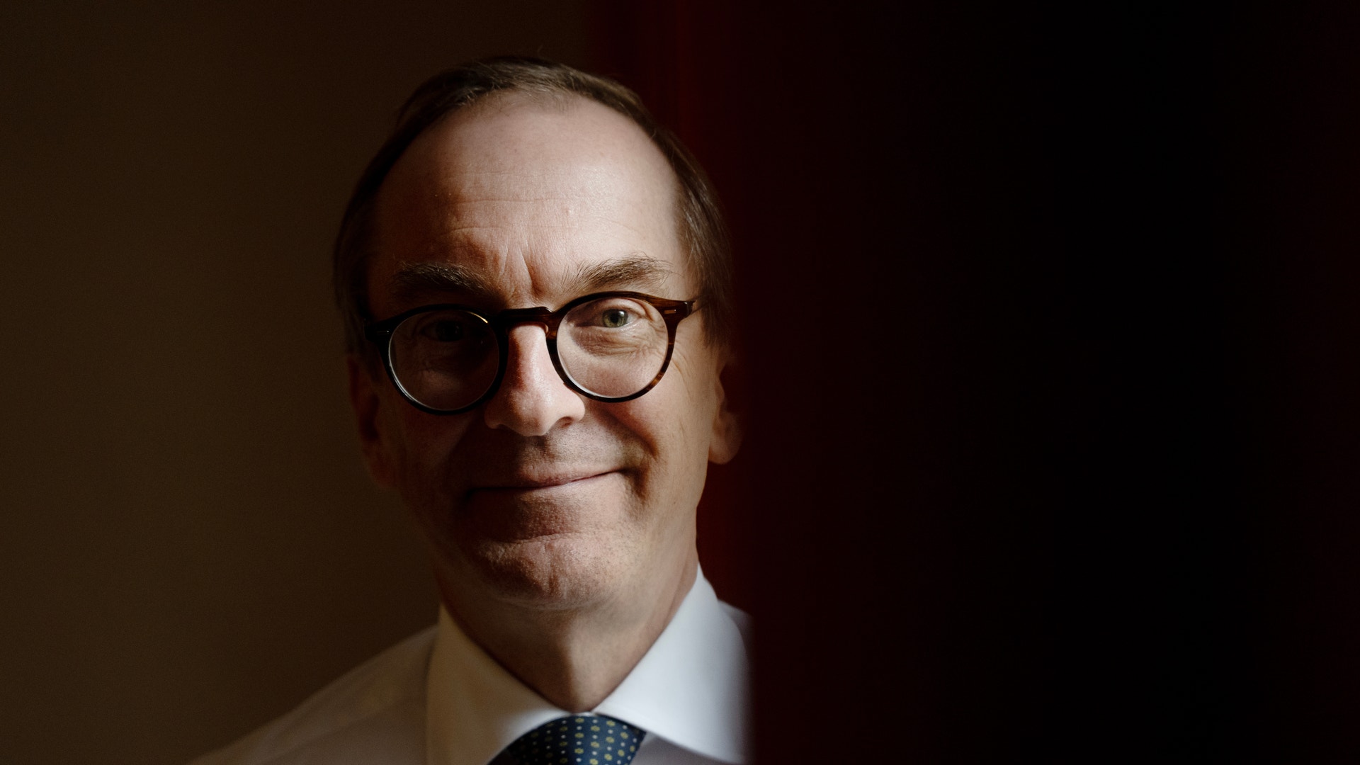 Porträtt av en man med glasögon, vit skjorta och mörkblå slips mot mörk bakgrund.