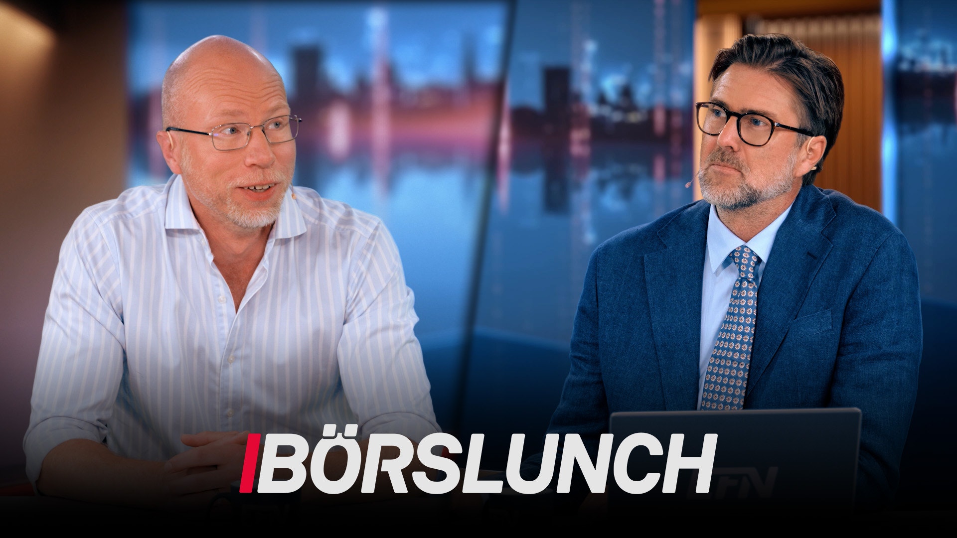 Två män i affärskläder sitter vid ett skrivbord under programtiteln Börslunch. I bakgrunden syns en stadshorisont.
