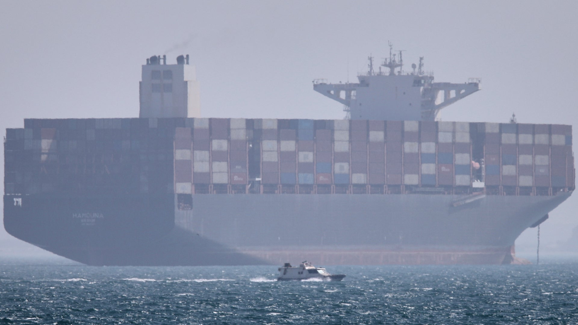 Ett stort containerfartyg på öppet hav med hundratals blå och grå containrar staplade på däck. En liten båt syns i förgrunden på det skummiga vattnet.