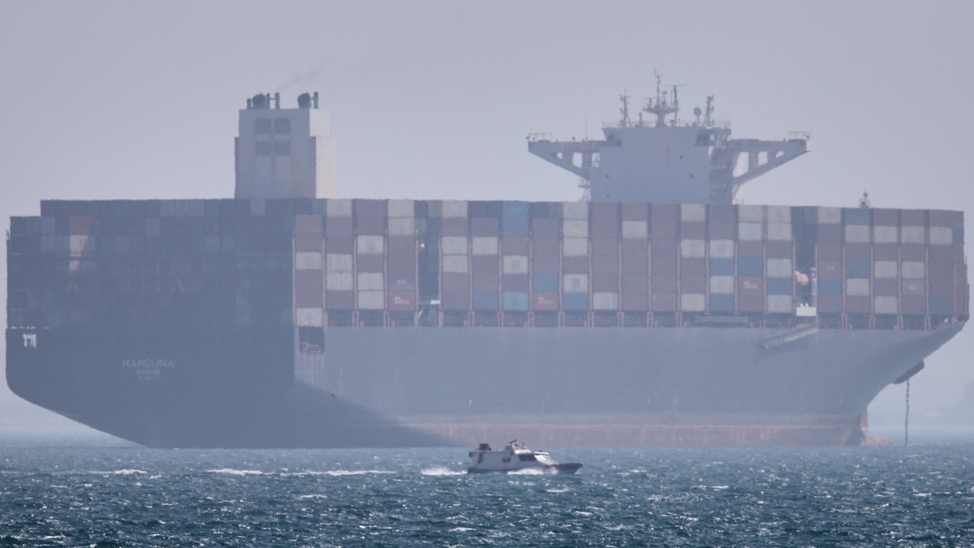 Ett stort containerfartyg på öppet hav med hundratals blå och grå containrar staplade på däck. En liten båt syns i förgrunden på det skummiga vattnet.