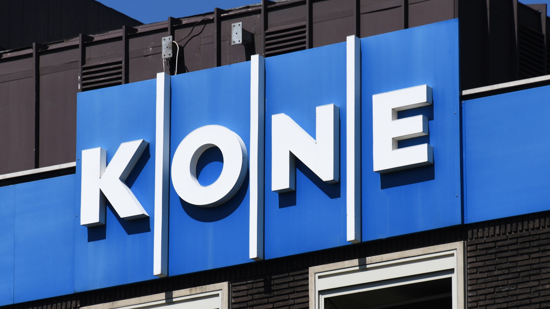 Blå skylt med vita bokstäver som visar texten "KONE" på en byggnadsfasad