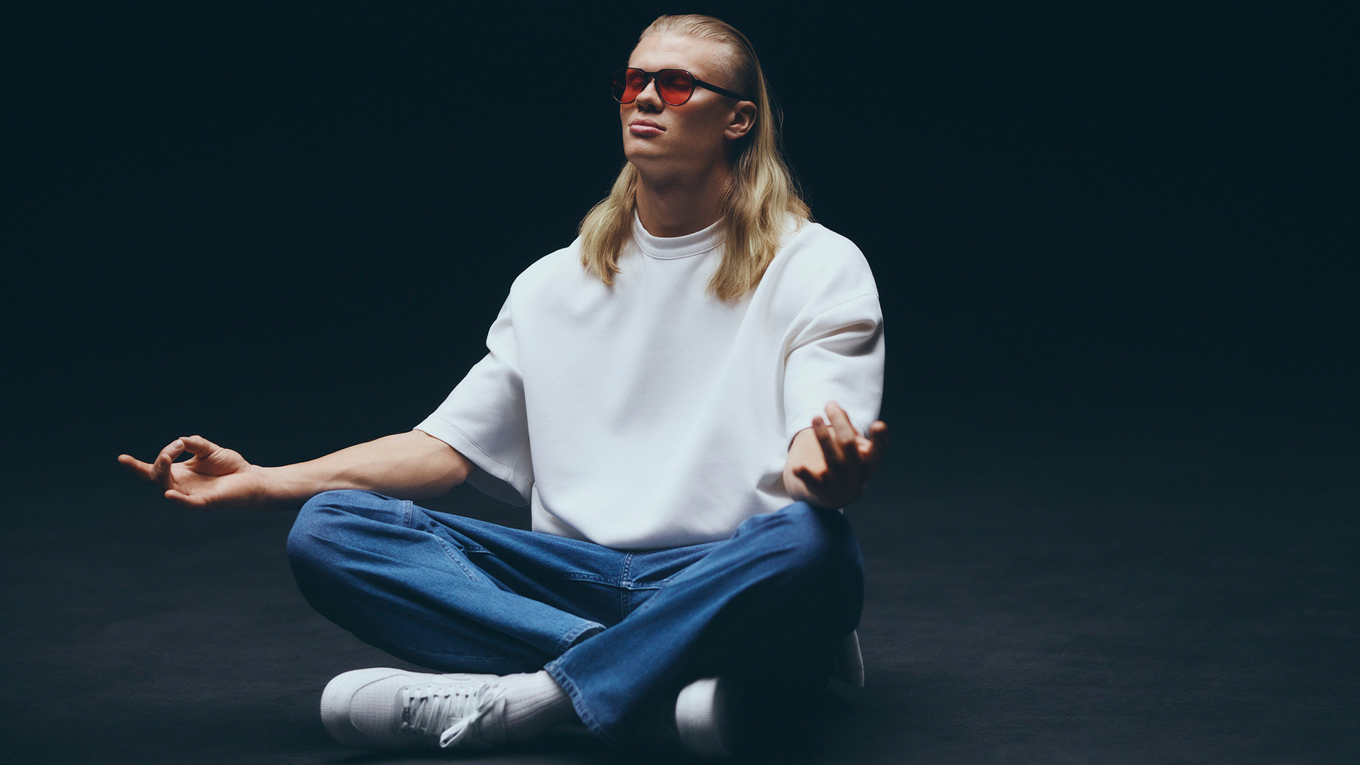 Person med långt blont hår sitter i lotusposition mot mörk bakgrund. Personen bär vita solglasögon, vit t-shirt och blå jeans med armarna utsträckta.