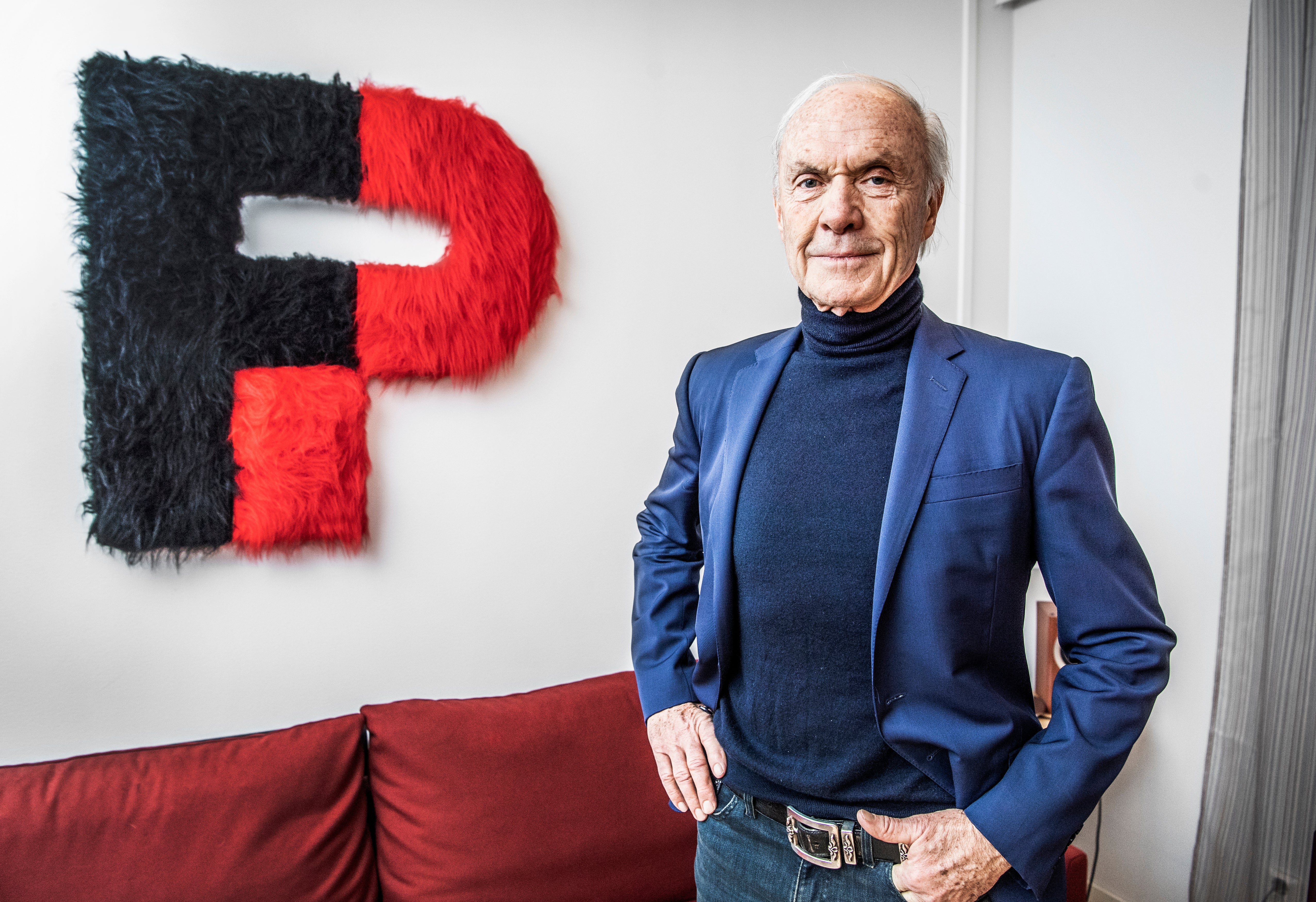 Porträttfoto av en äldre man i blå kavaj som står framför en röd och svart logotyp på vit vägg.