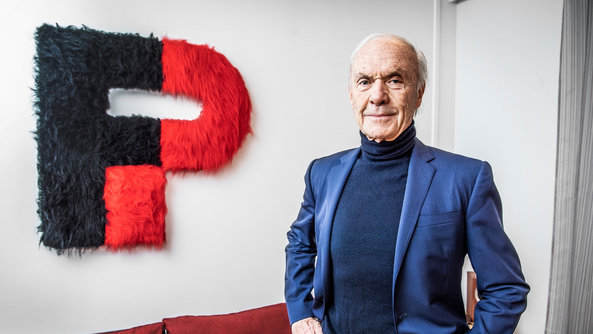 Porträttfoto av en äldre man i blå kavaj som står framför en röd och svart logotyp på vit vägg.