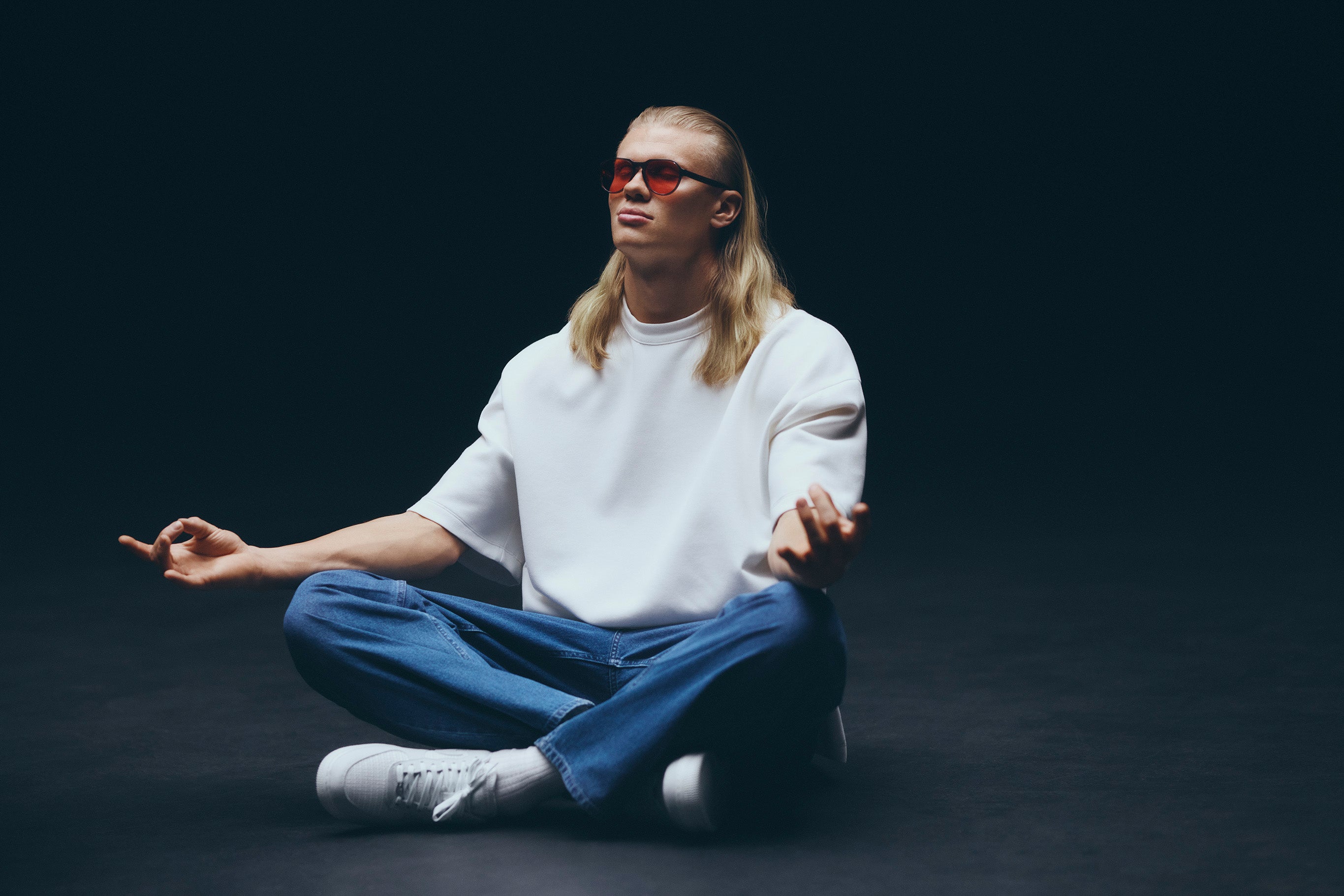 Person med långt blont hår och glasögon sitter på golvet mot mörk bakgrund. Personen bär vit t-shirt och blå jeans.