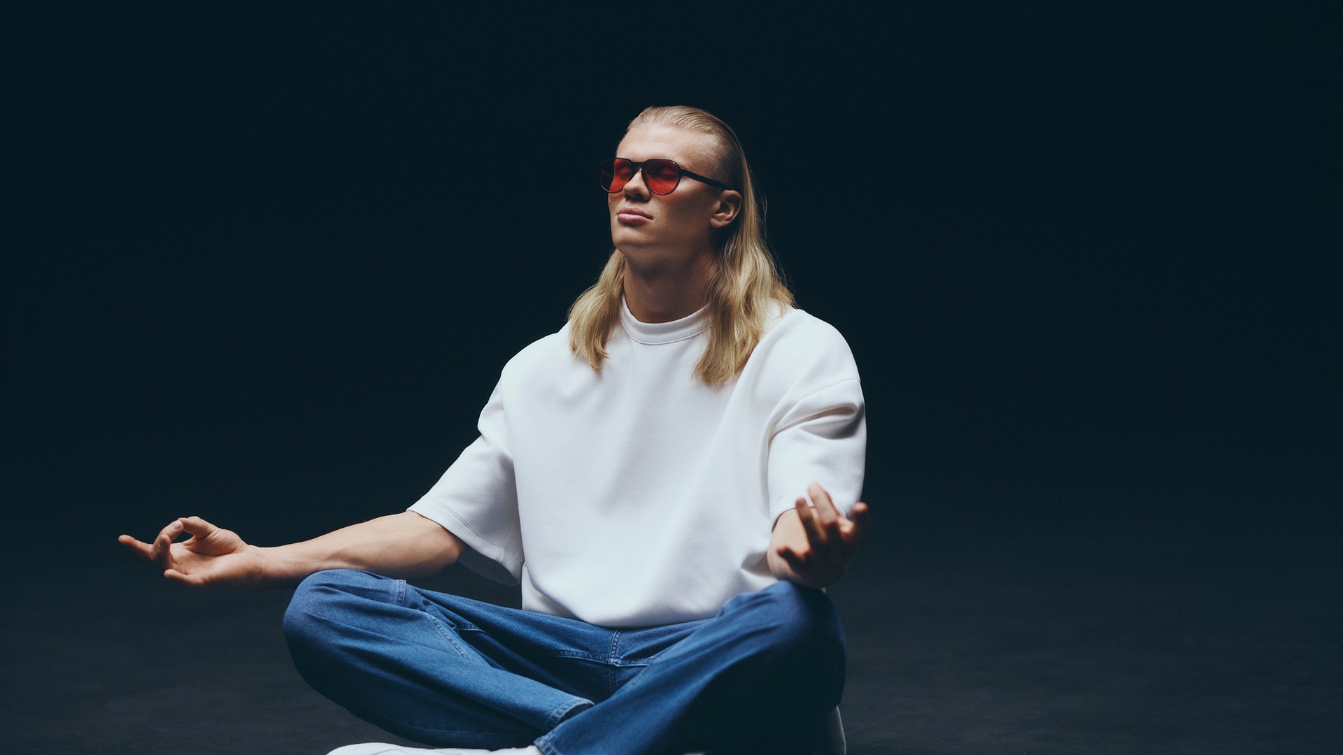 Person med långt blont hår och glasögon sitter på golvet mot mörk bakgrund. Personen bär vit t-shirt och blå jeans.
