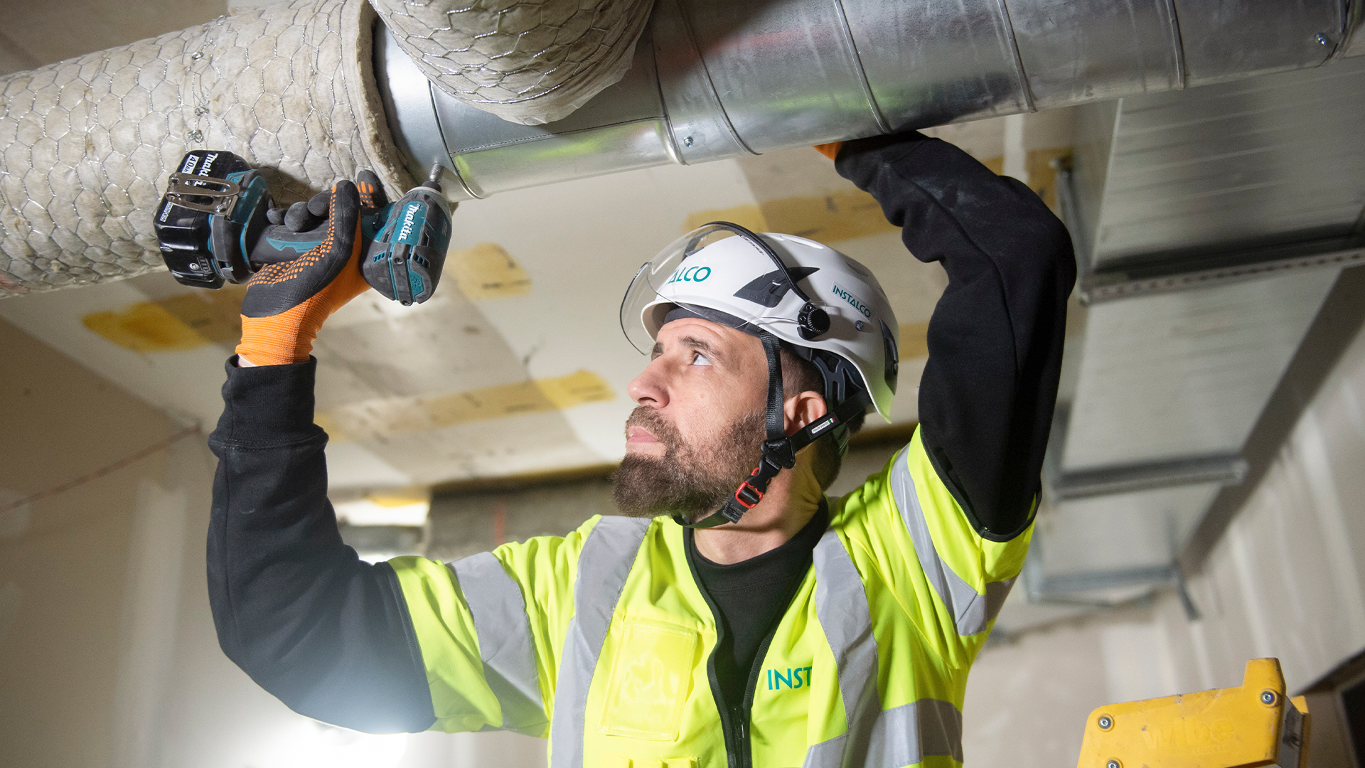 Arbetare i gul väst och skyddshjälm installerar ventilationsrör i ett tak.