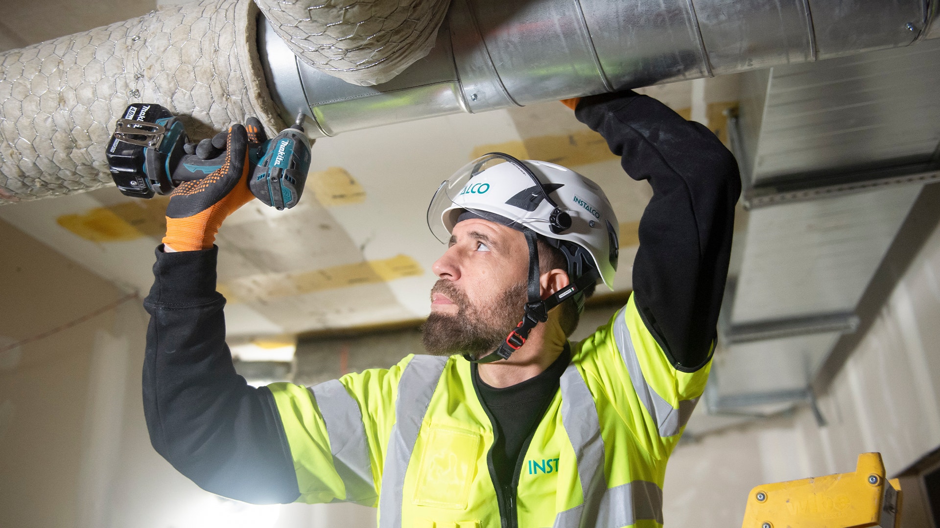 Arbetare i gul väst och skyddshjälm installerar ventilationsrör i ett tak.