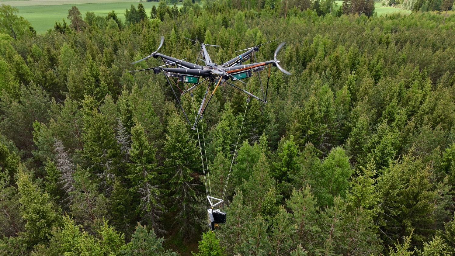 En drönare flyger över en grön skog fylld med barrträd. Drönaren är utrustad med fyra rotorblad och bär utrustning under sig.