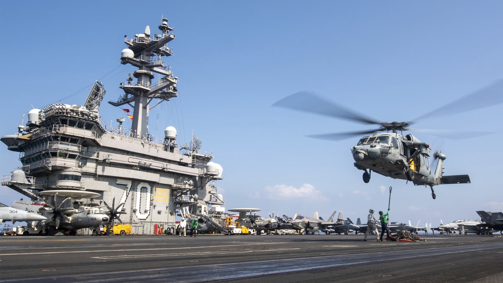 En militär helikopter landar på ett hangarfartygs flydäck. Fartyget med sina höga master och antenner syns i bakgrunden, medan helikoptern är i luften med rotorbladen i rörelse.