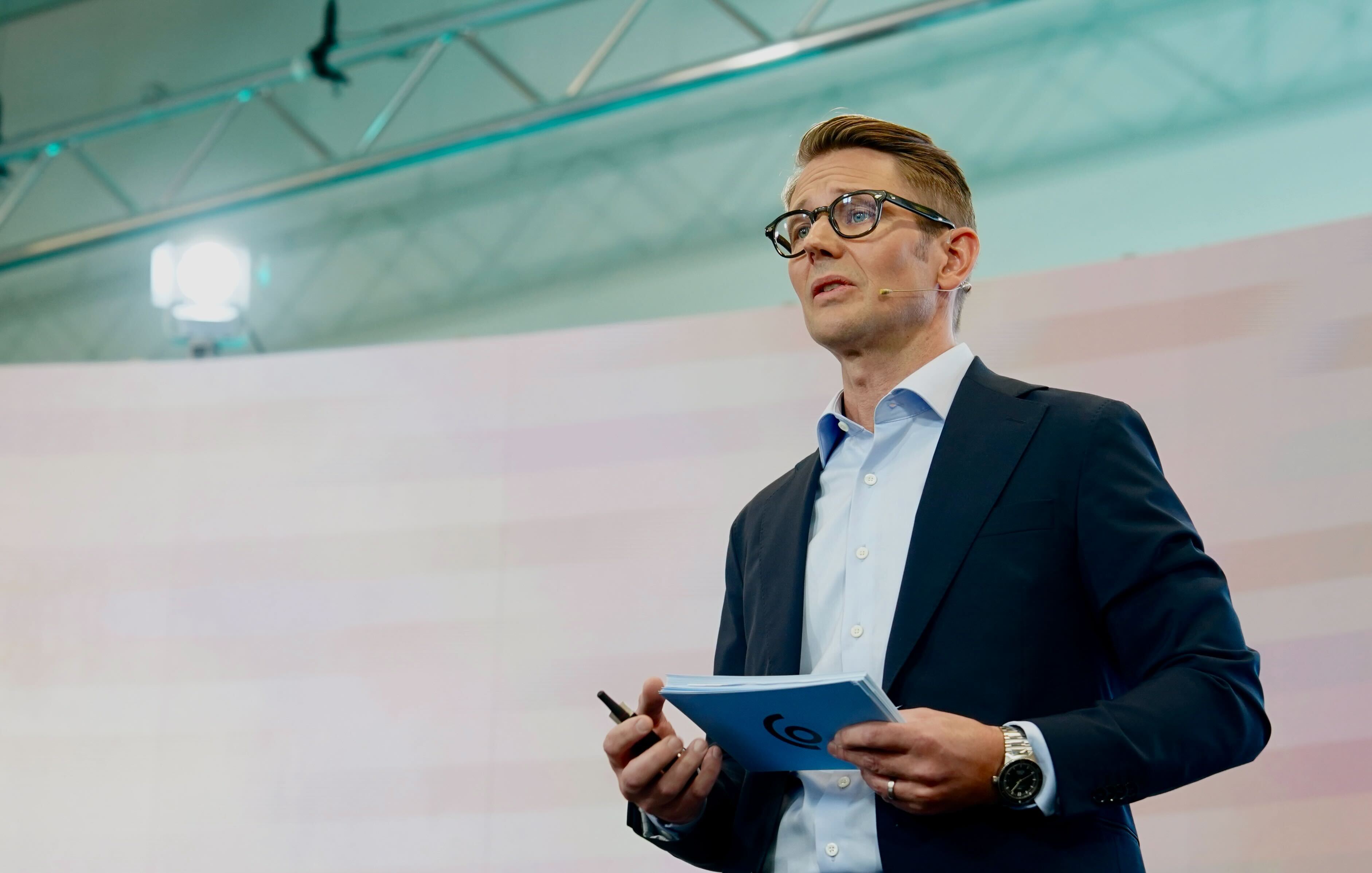 Man i kostym och glasögon presenterar med en tablet framför en projektionsskärm i en modern sal.
