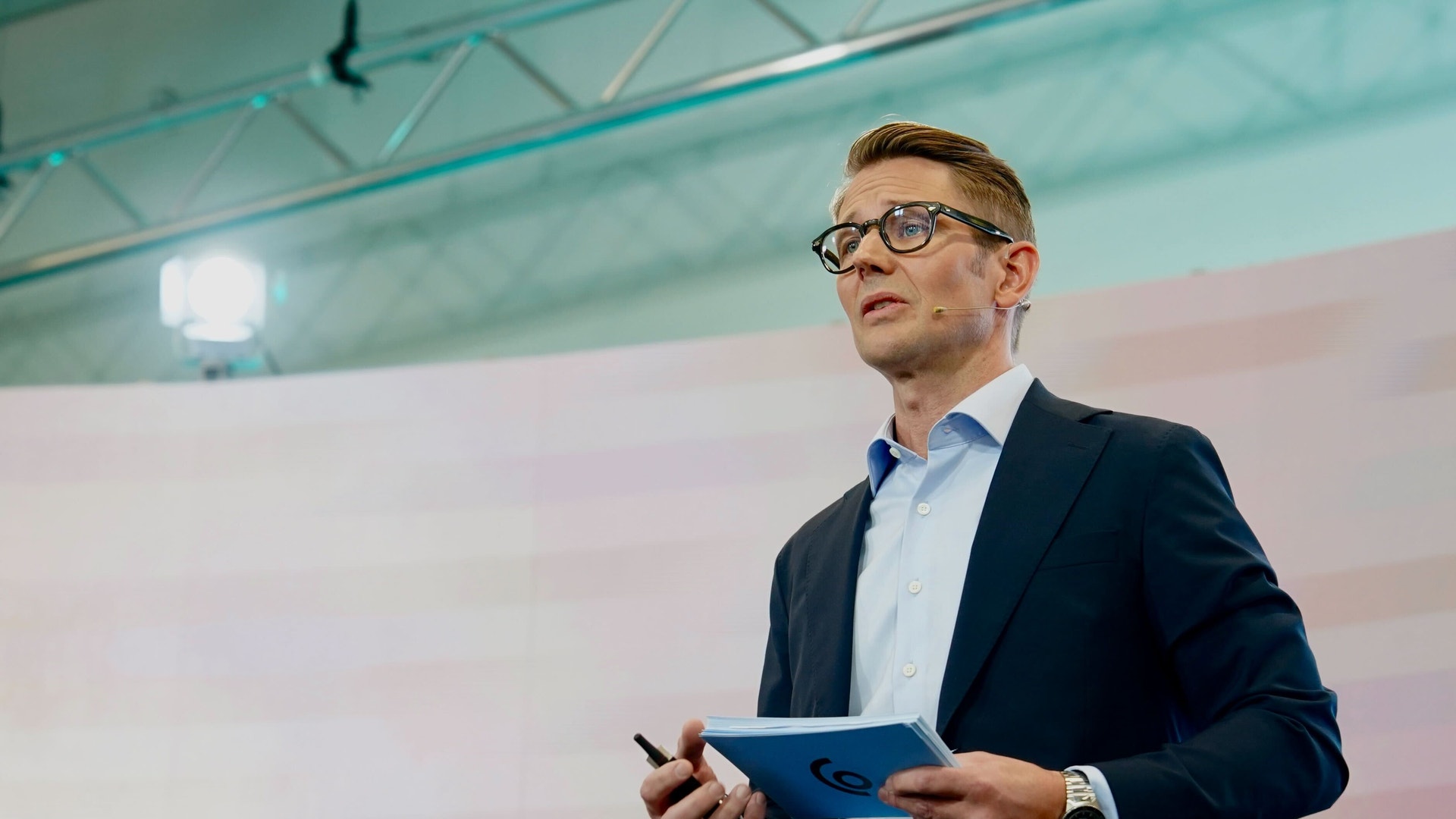 Man i kostym och glasögon presenterar med en tablet framför en projektionsskärm i en modern sal.