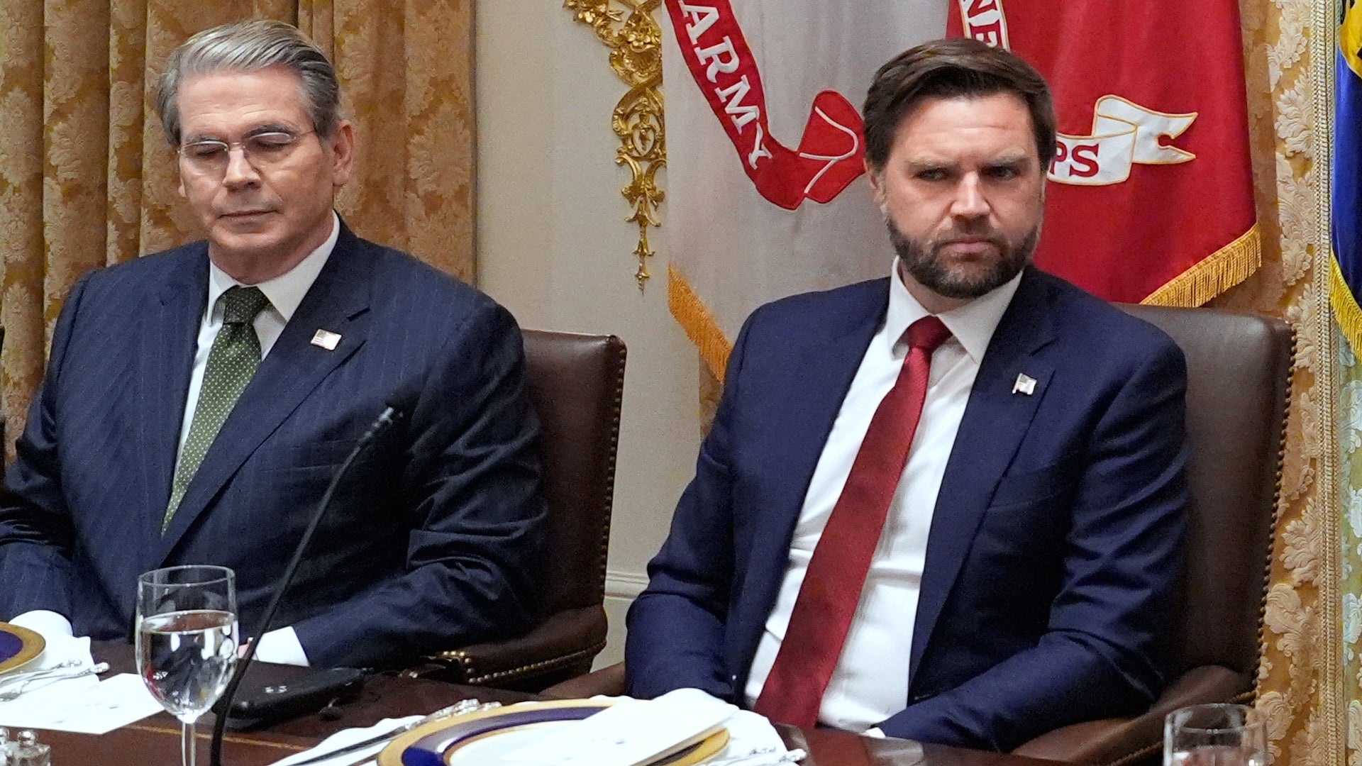 Två män i kostym sitter vid ett formellt mötesbord med röda flaggor och officiella emblem i bakgrunden.