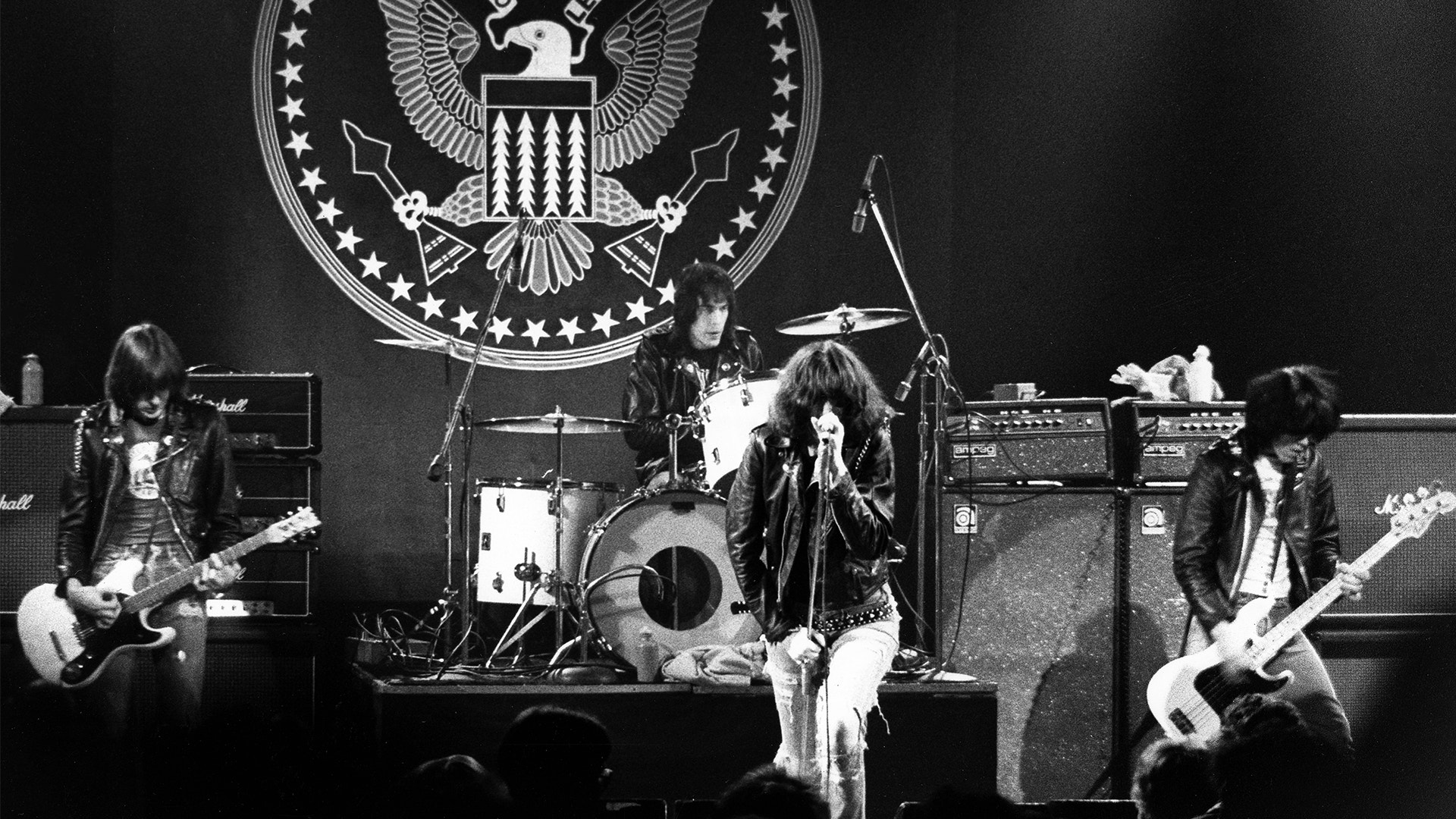 Svartvit fotografi av rockband som framför live på scen med gitarrister, trummis och sångare framför en stor örn-emblem.