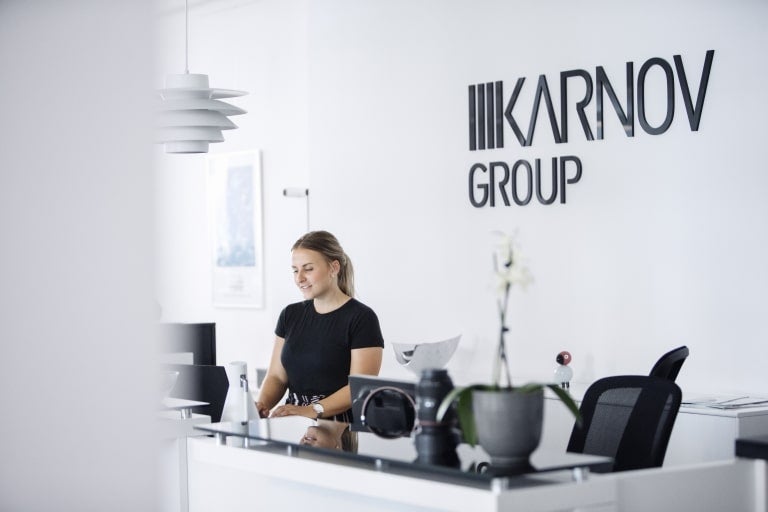 Receptionist sitter vid ett vitt skrivbord i ett modernt kontor med Karnov Group-logotypen på väggen bakom.