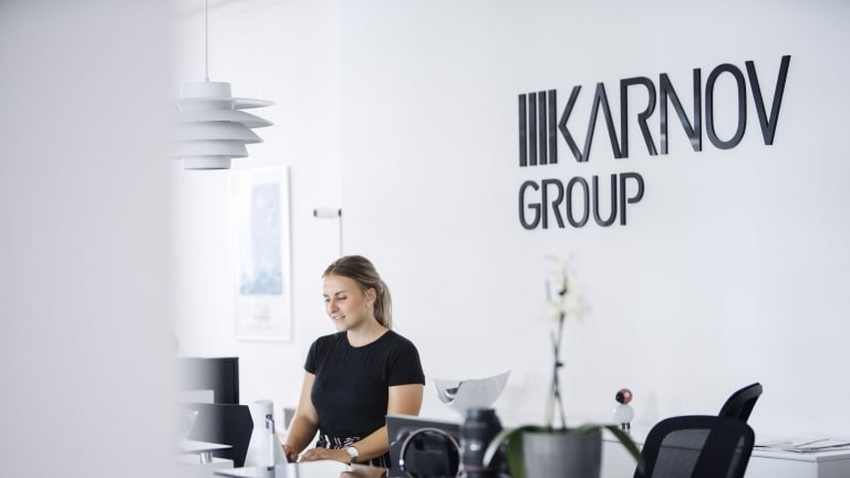 Receptionist sitter vid ett vitt skrivbord i ett modernt kontor med Karnov Group-logotypen på väggen bakom.
