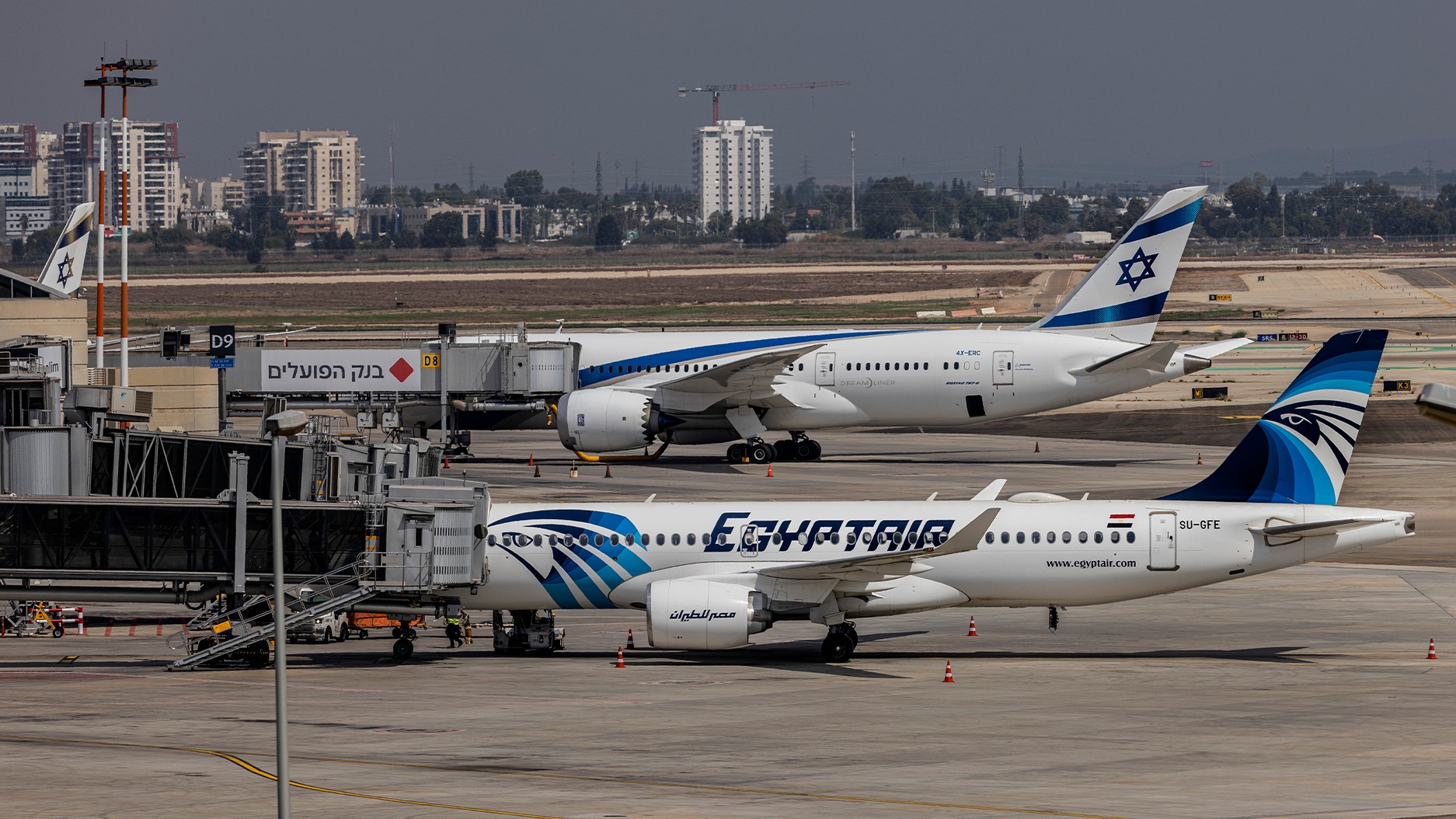 Två passagerarflygplan på en flygplats. I förgrunden en EgyptAir-maskin och bakom den ett El Al-plan. Flygplatsens byggnader och stadssiluett syns i bakgrunden.