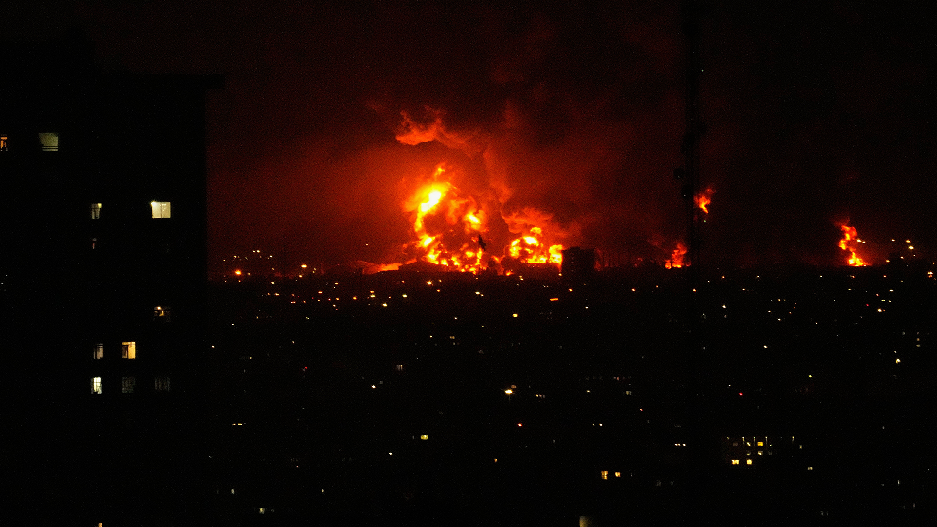 Nattlig stadsbild med stora bränder som lyser upp himlen i orange och gult ljus