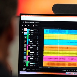 Skärmdump av musikproduktionsprogram SUND Studio med flera färgglada ljudspår visade som vågformer på en datorskärm. En webbkamera är monterad ovanpå skärmen.