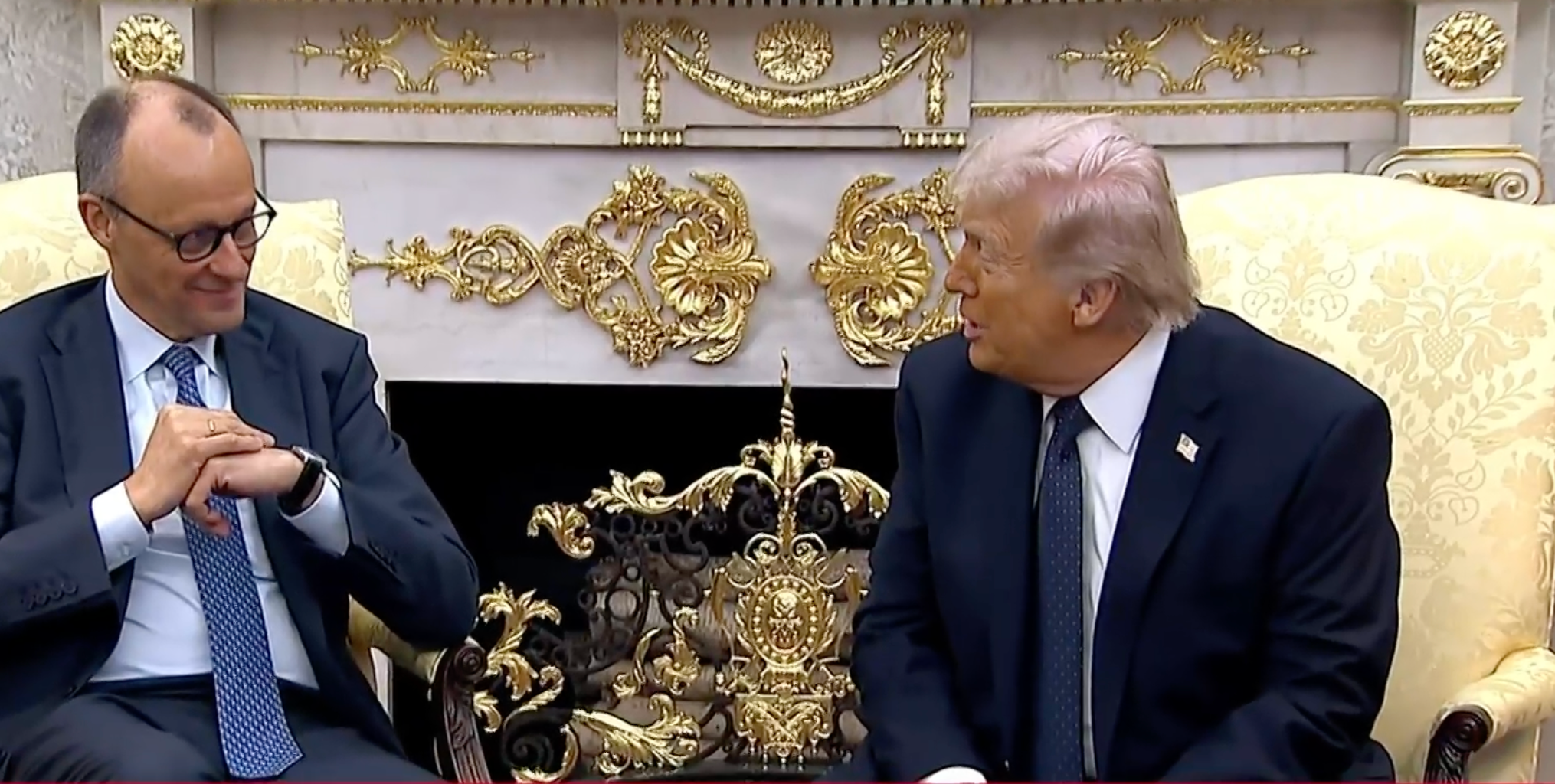 Två män i kostym sitter i en prunkig inredning med gulddekorationer och samtalar med varandra.