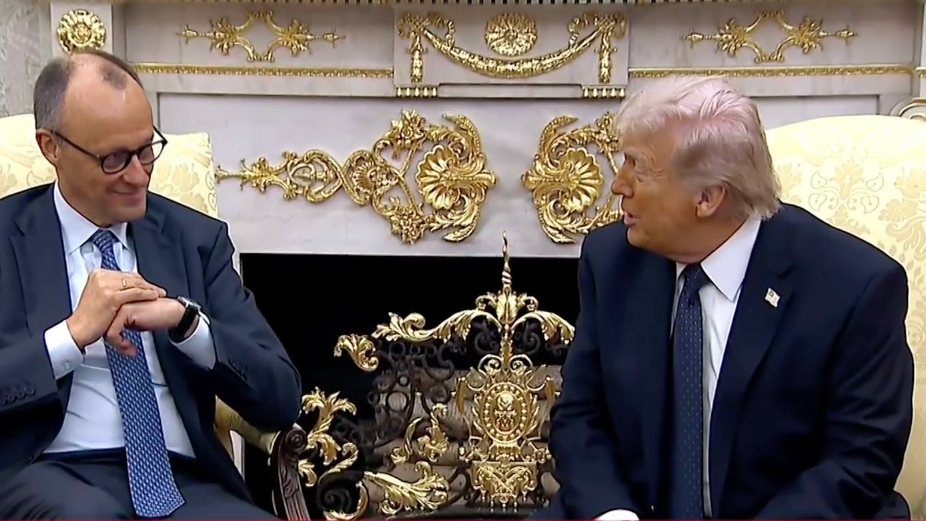 Två män i kostym sitter i en prunkig inredning med gulddekorationer och samtalar med varandra.