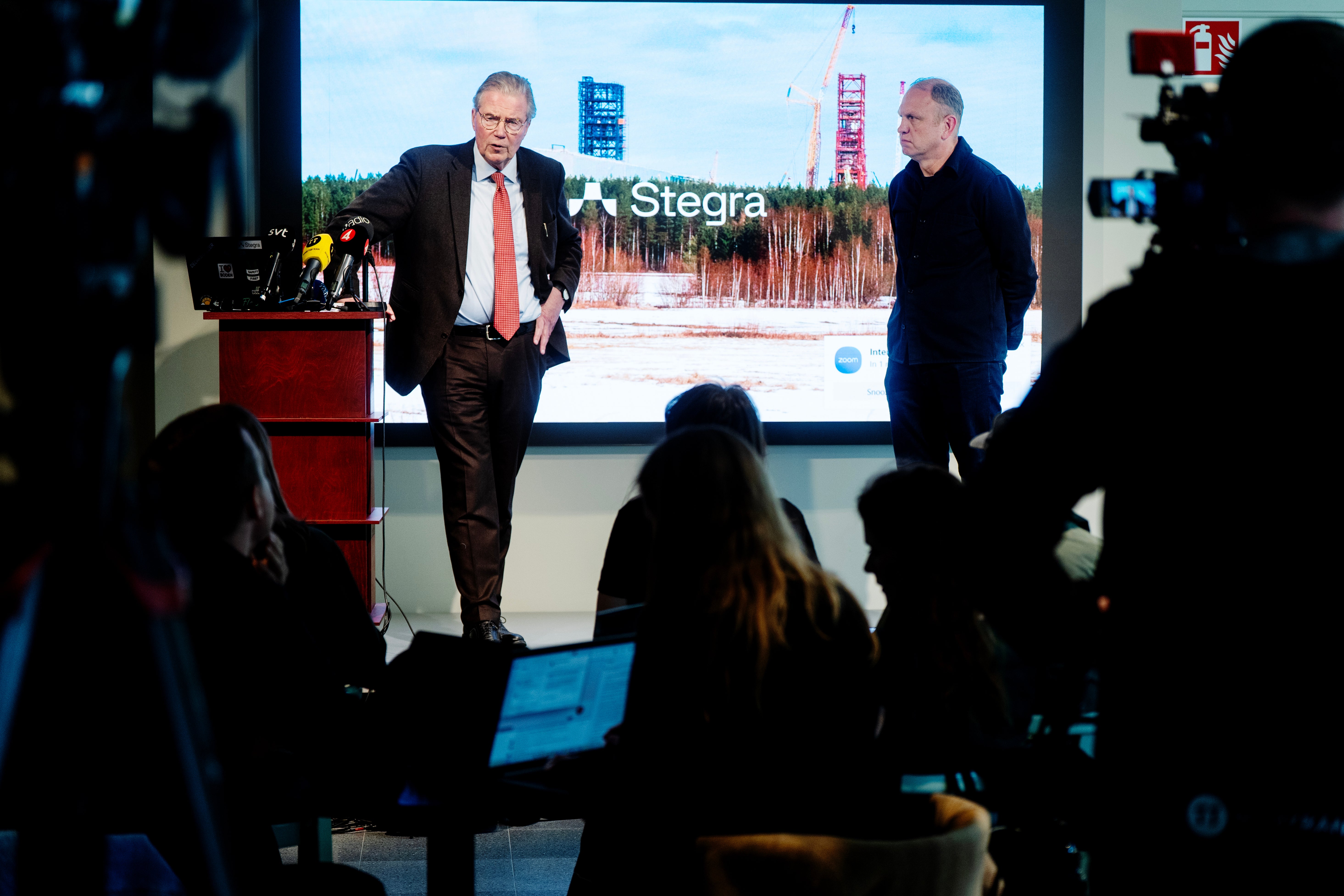 Två män presenterar på en scen framför publik med en stor skärm bakom sig som visar Stegra-logotypen och en industriell anläggning.
