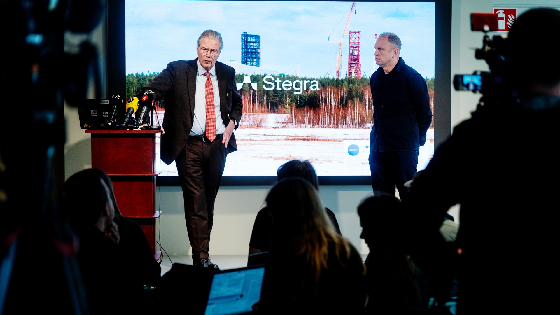 Två män presenterar på en scen framför publik med en stor skärm bakom sig som visar Stegra-logotypen och en industriell anläggning.