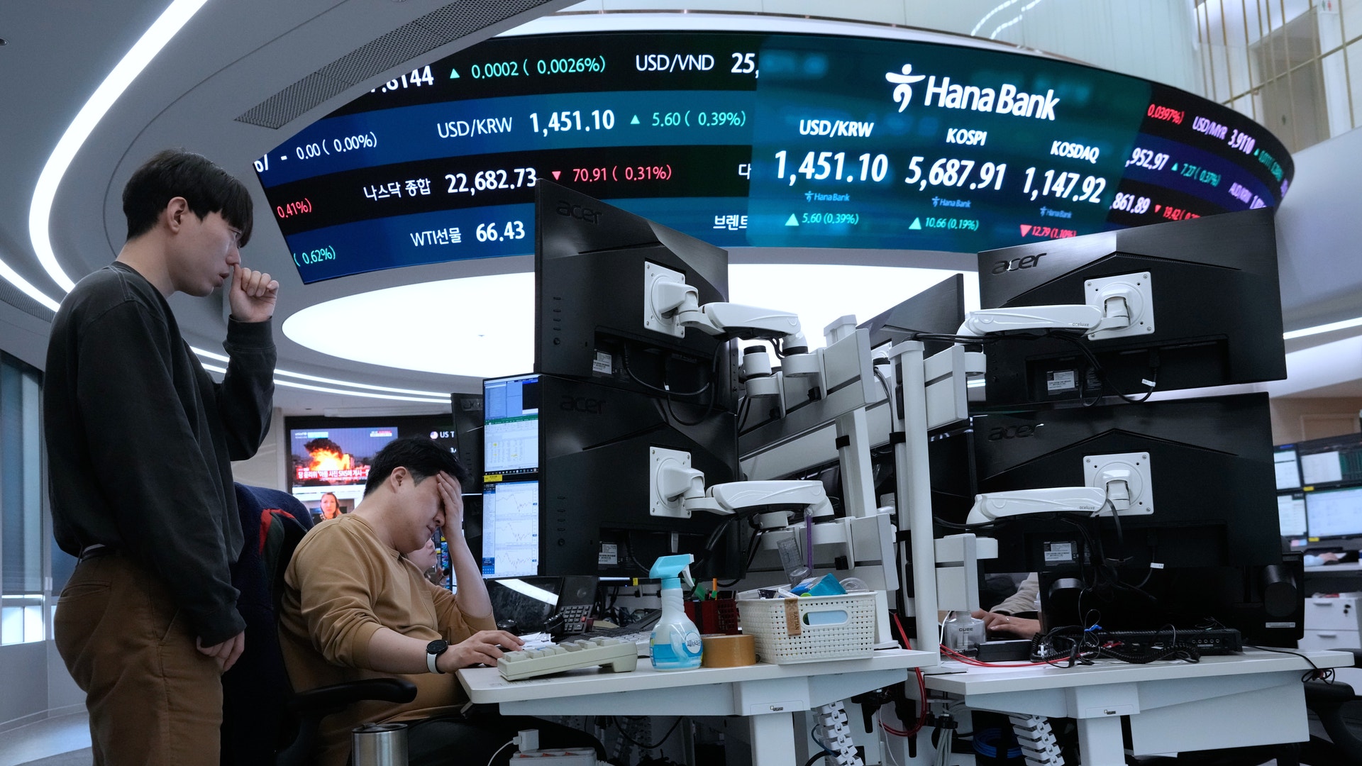 Två personer arbetar vid ett handelsskrivbord med finansiella marknadsöversikter och aktiekurser visade på stora skärmar ovanför.