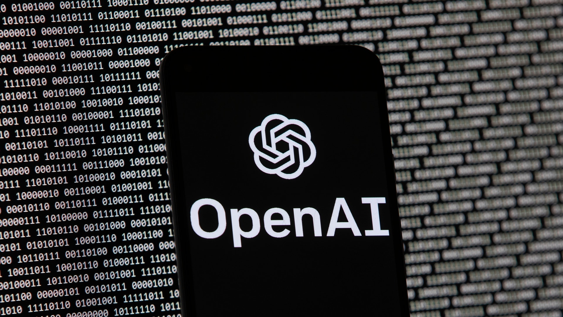 En mobiltelefon visar OpenAI:s logotyp mot en bakgrund med binär kod.