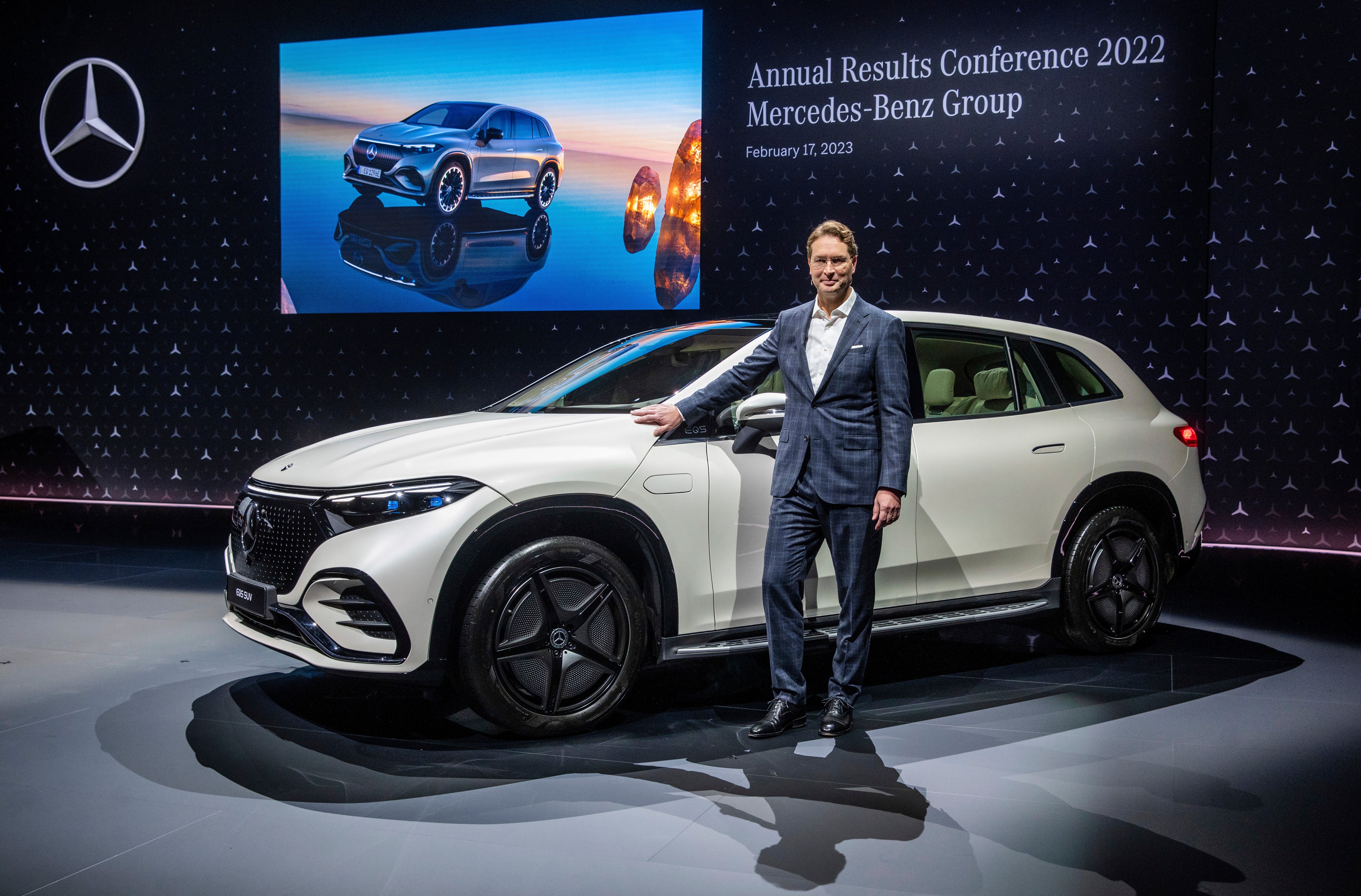 En man i kostym står bredvid en vit Mercedes-Benz-bil på en scen under en företagspresentation.