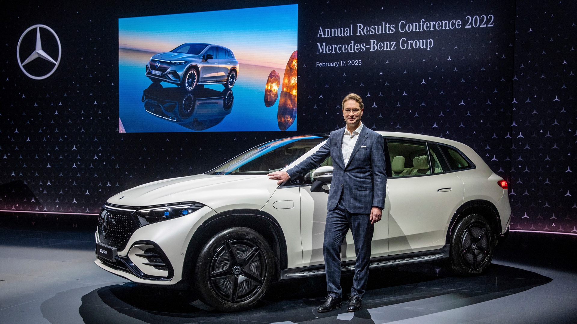 En man i kostym står bredvid en vit Mercedes-Benz-bil på en scen under en företagspresentation.