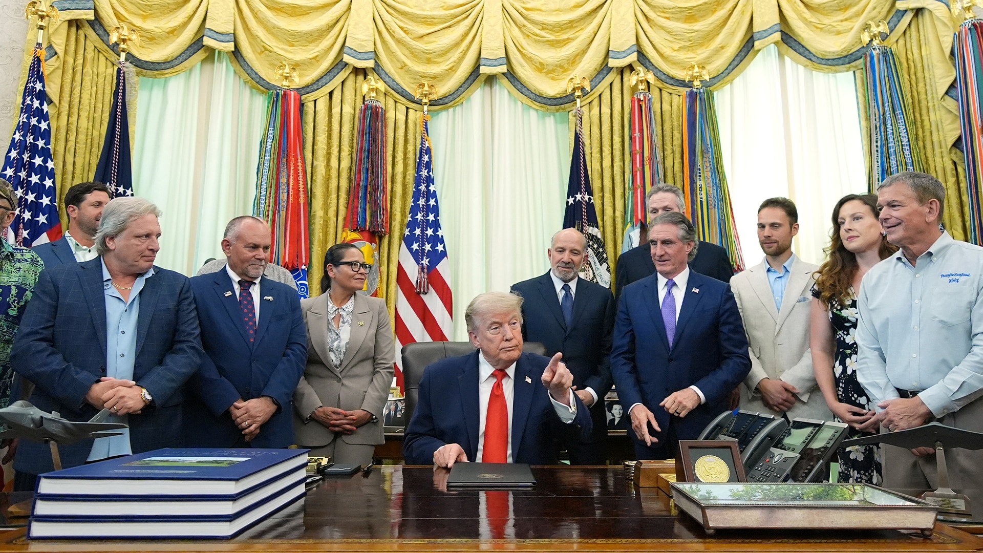 En grupp människor står omkring ett skrivbord i ett officiellt kontor. I bakgrunden syns amerikanska flaggor och gula gardiner.
