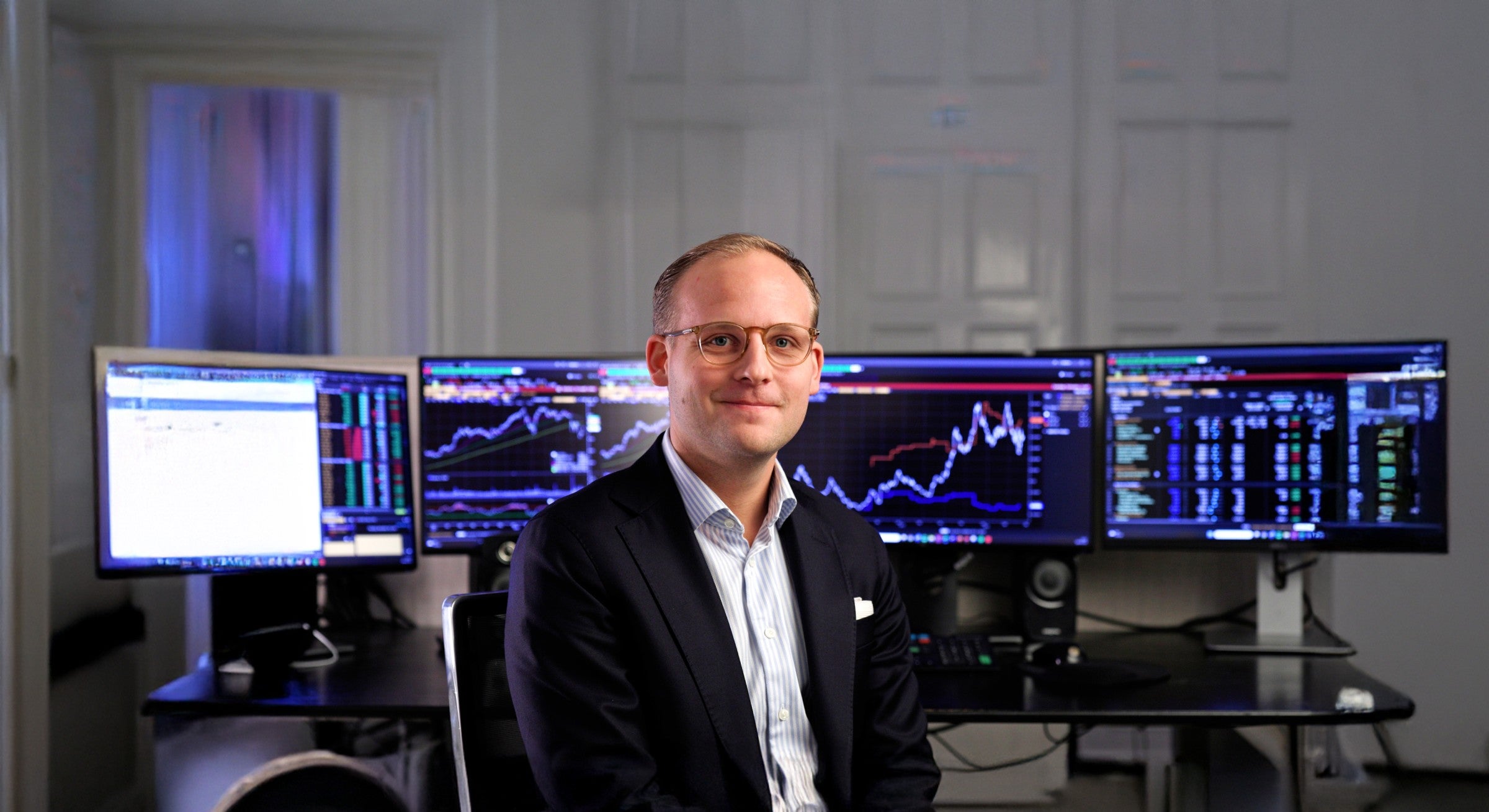 Man i glasögon och kostym sitter framför flera datorskärmar med finansiella grafer och marknadsdata.
