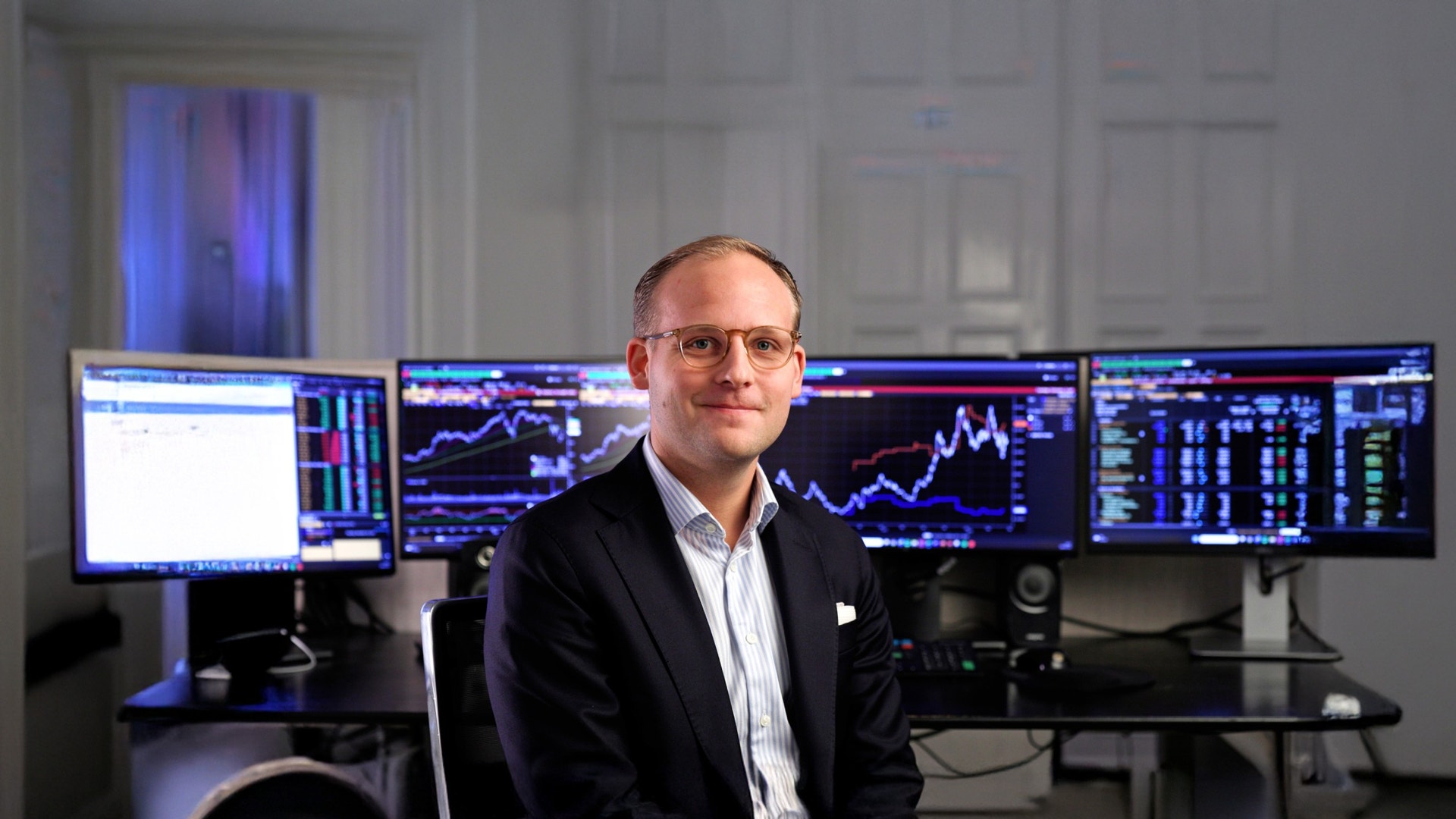 Man i glasögon och kostym sitter framför flera datorskärmar med finansiella grafer och marknadsdata.