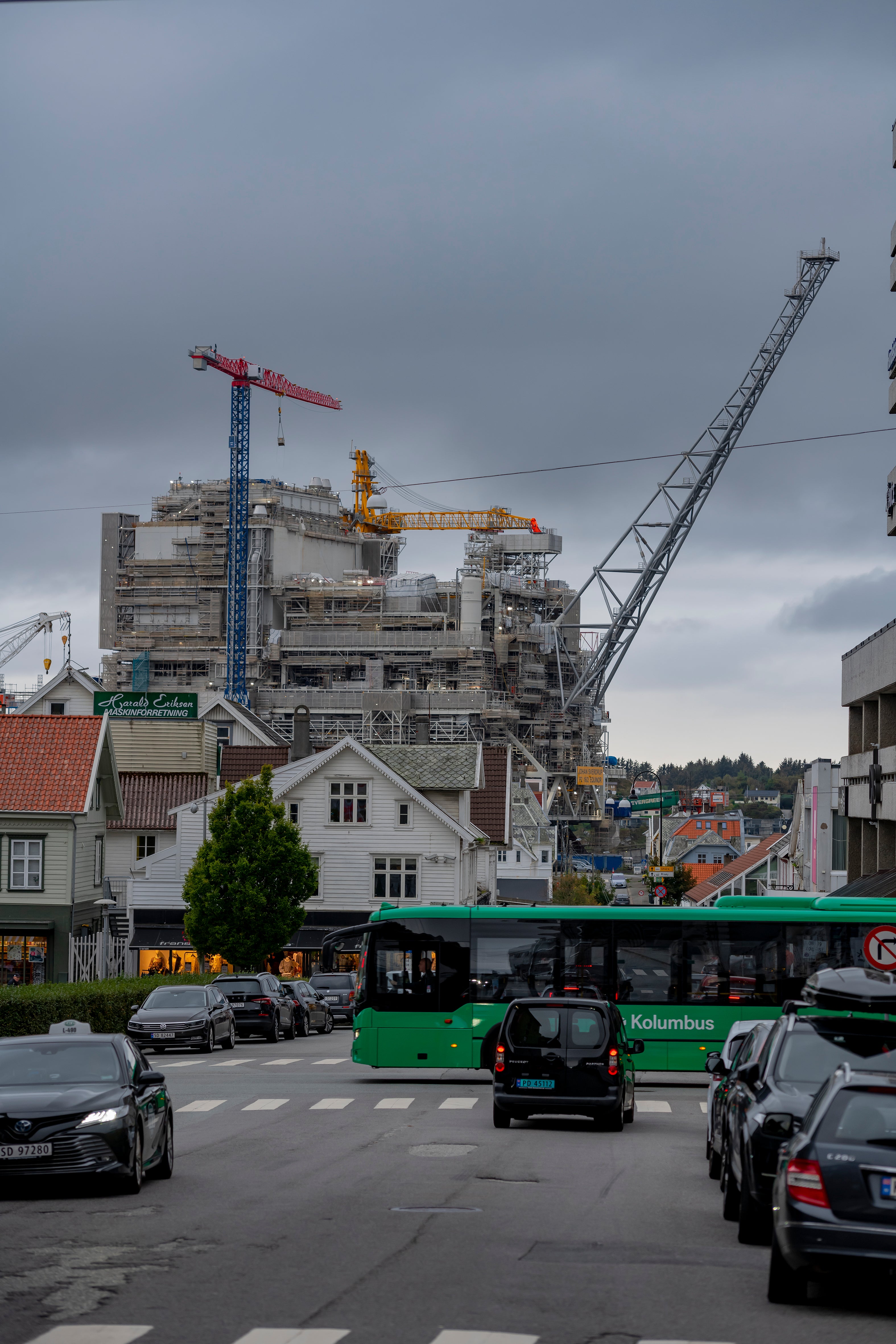 Industriell plattform under konstruktion med stora kranar och byggnadsställningar som reser sig över bostadshus och gata med parkerade bilar.