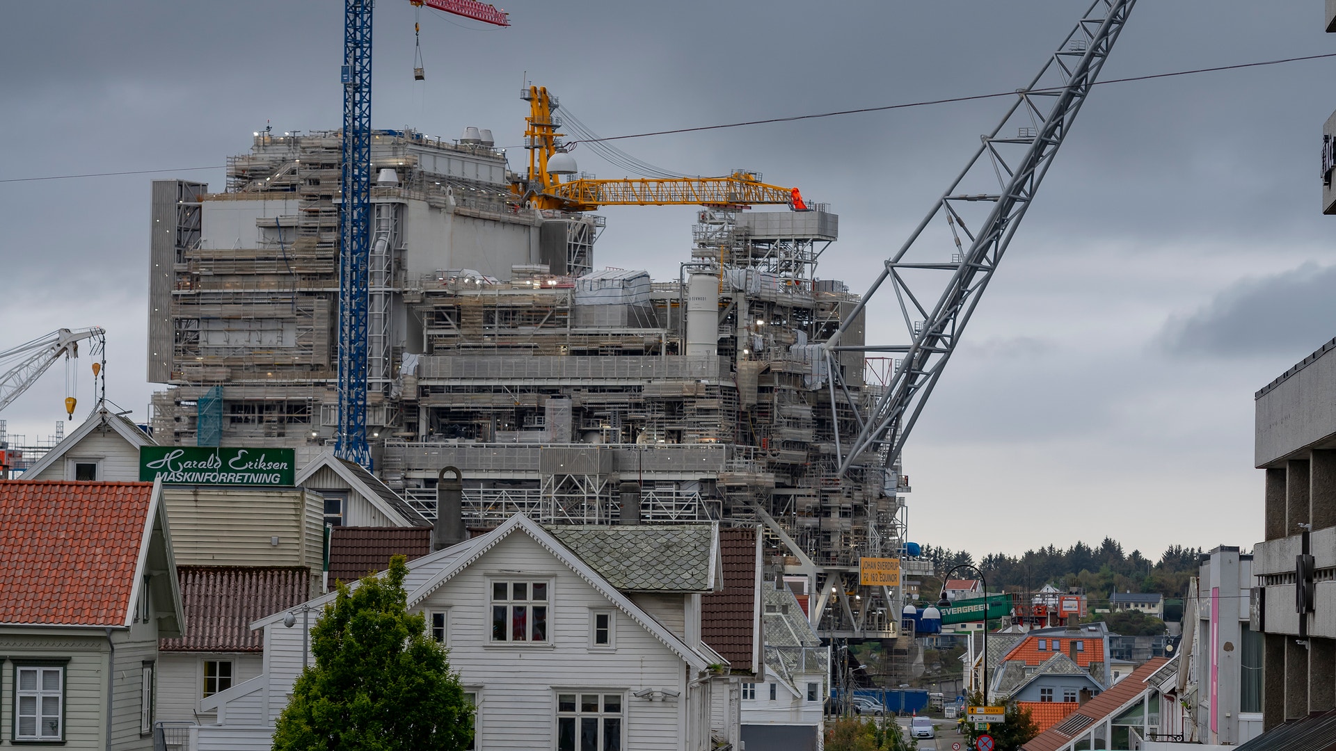 Industriell plattform under konstruktion med stora kranar och byggnadsställningar som reser sig över bostadshus och gata med parkerade bilar.