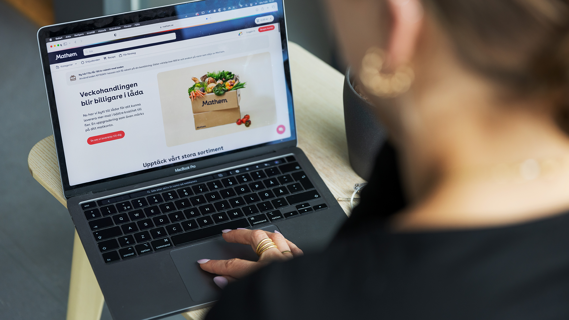 En person använder en laptop för att handla mat online på en matvaruwebbshop. Skärmen visar en webbutik med en bild på en matkasse med grönsaker och andra livsmedel.
