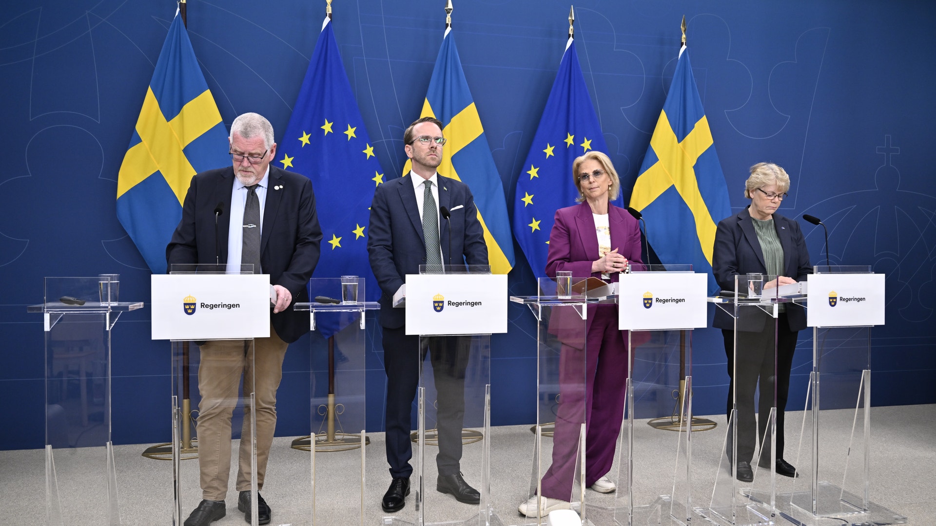 Fyra personer står vid podier under en presskonferens. I bakgrunden syns svenska flaggor och EU-flaggor på en blå vägg.