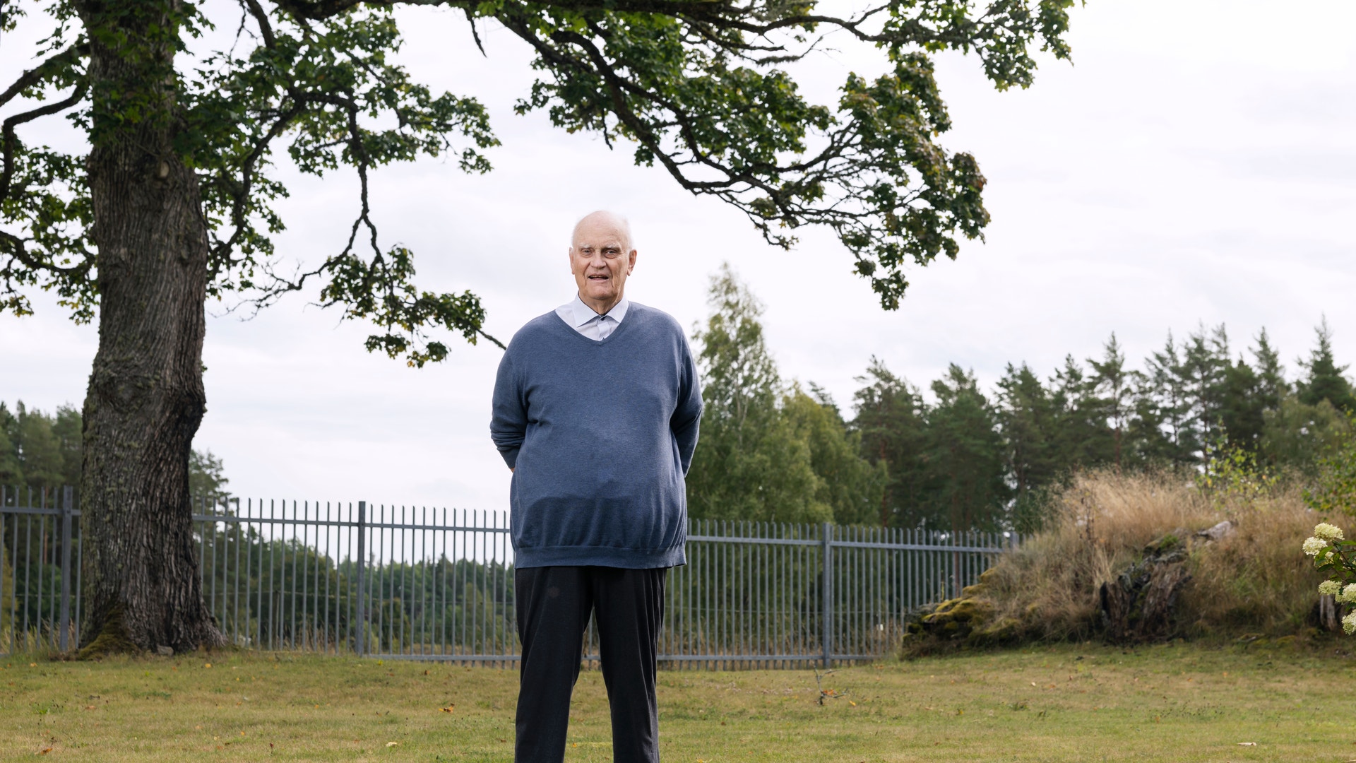 En äldre man står på en gräsmatta framför ett stort träd. Han bär en blå tröja och mörkblå byxor. I bakgrunden syns ett staket och gröna träd.