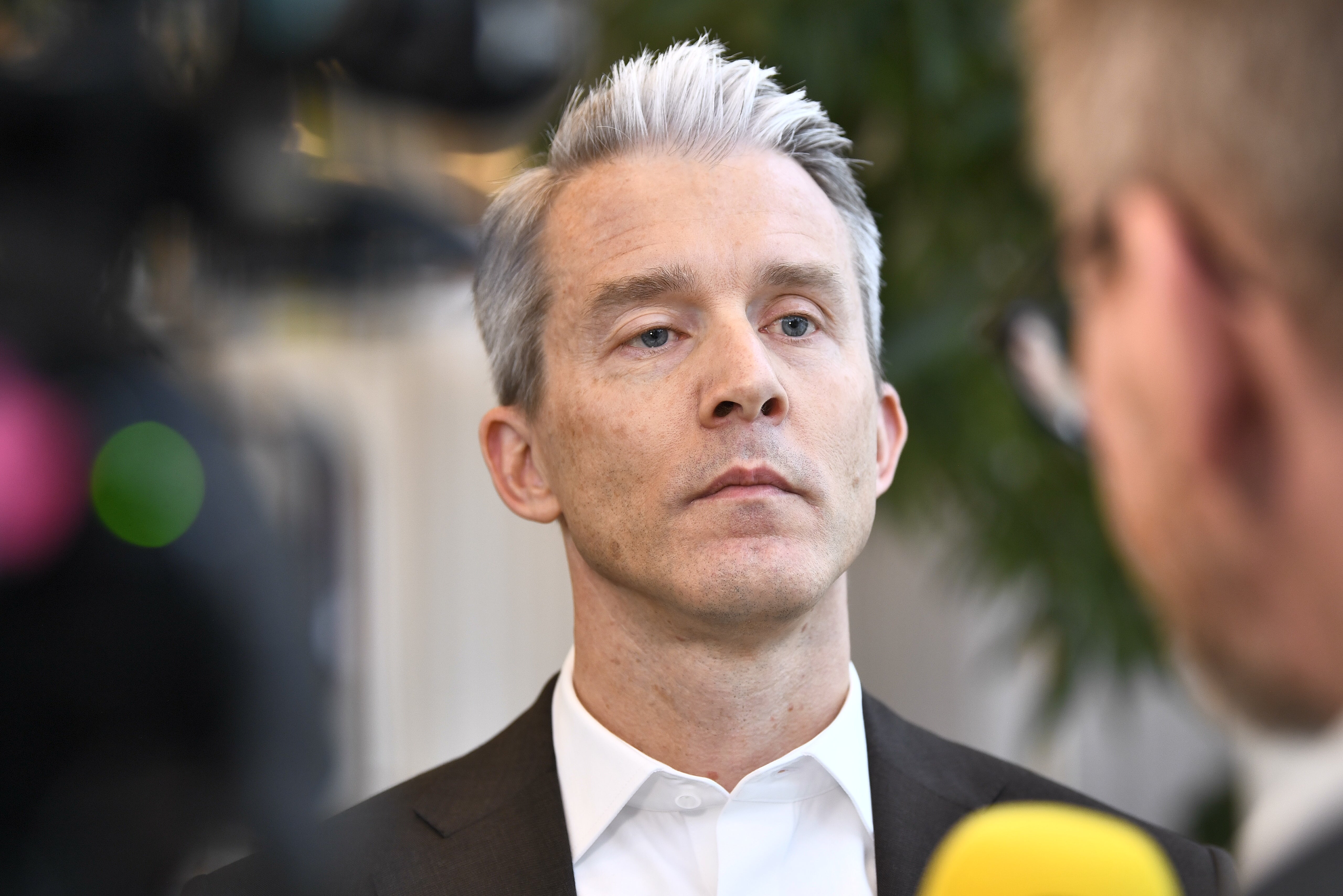 Porträtt av en man med grått hår och mörk kavaj som tittar allvarligt åt sidan, med mikrofoner framför sig.