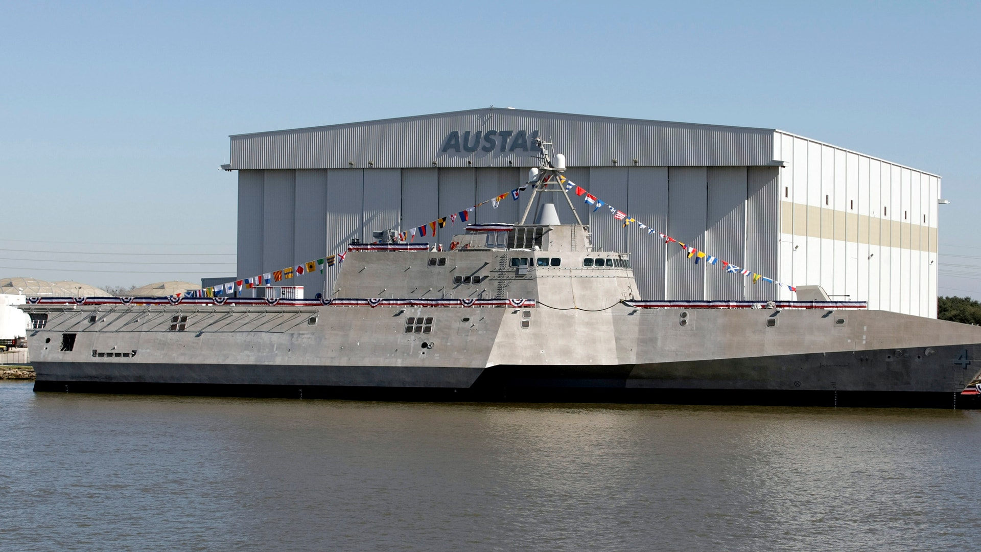 Ett militärt krigsfartyg med grå skrov ligger dockat vid en industribyggnad. Fartyget är dekorerat med färgglada flaggor längs sidan.