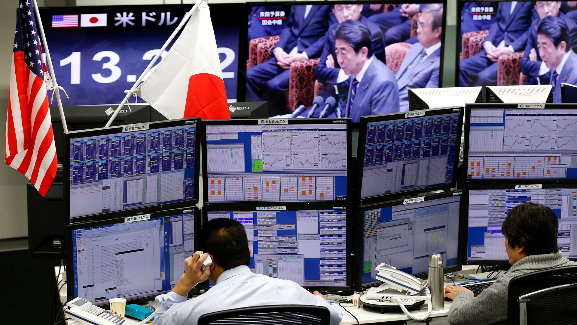 Person som arbetar vid ett handelsrum med flera datorskärmar som visar finansiell data och marknadsgrafer. Amerikanska och japanska flaggor syns i bakgrunden.