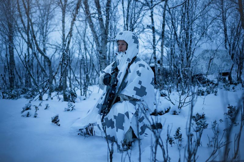 En soldat i vit snökamouflagekläder med gevär står i ett snötäckt skogslandskap under vinter.